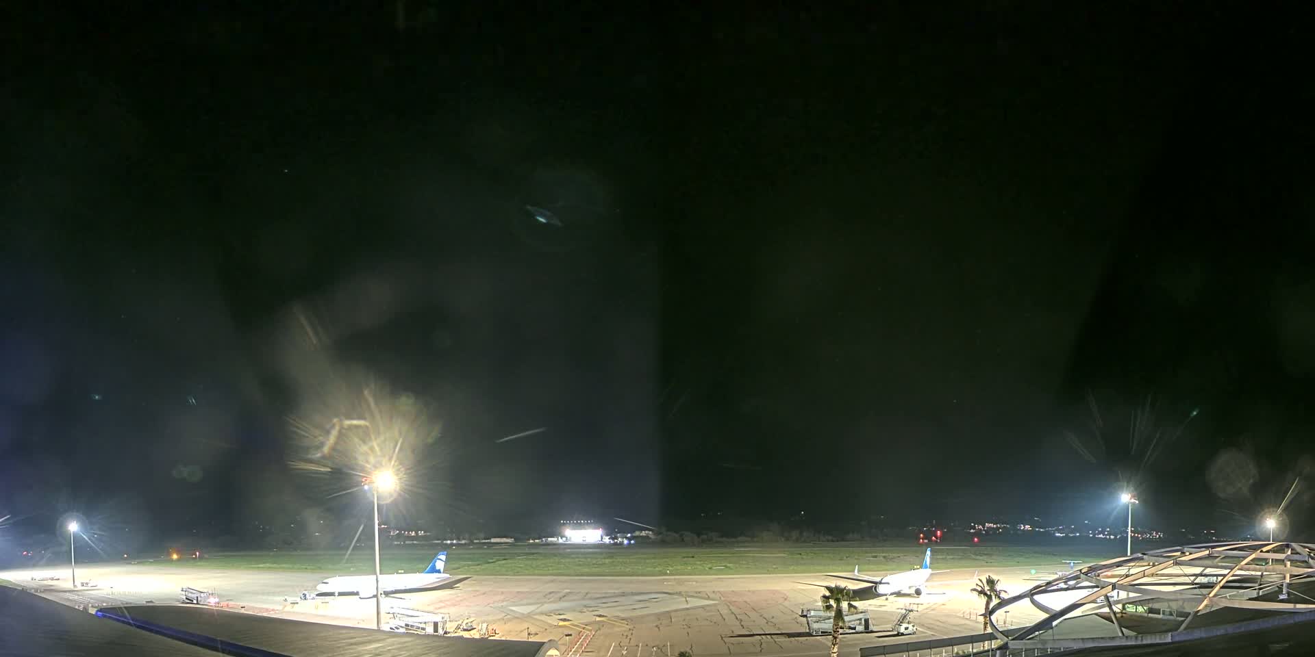 A nighttime view of an airport tarmac with two airplanes parked near gates, illuminated by bright lights under a dark, possibly rainy sky.