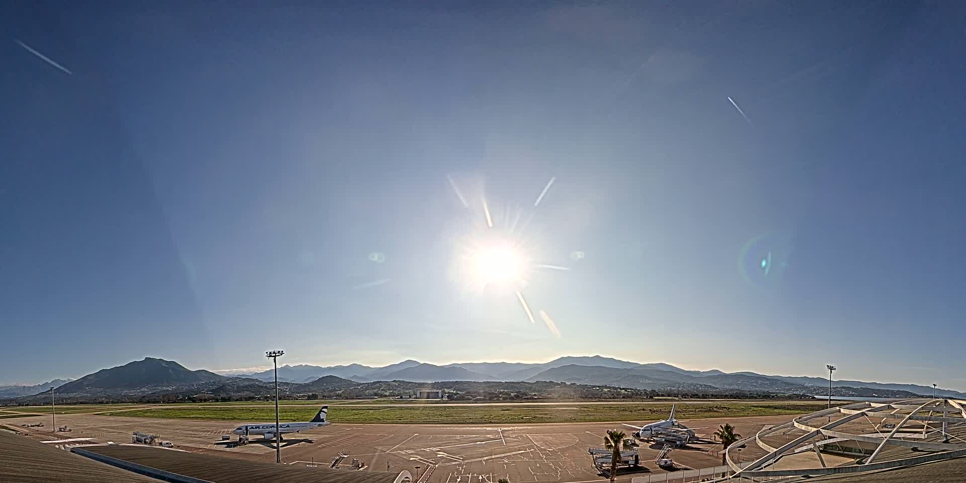 A bright, sunny day at an airport with two airplanes parked on the tarmac, overlooking a mountain range in the distance.