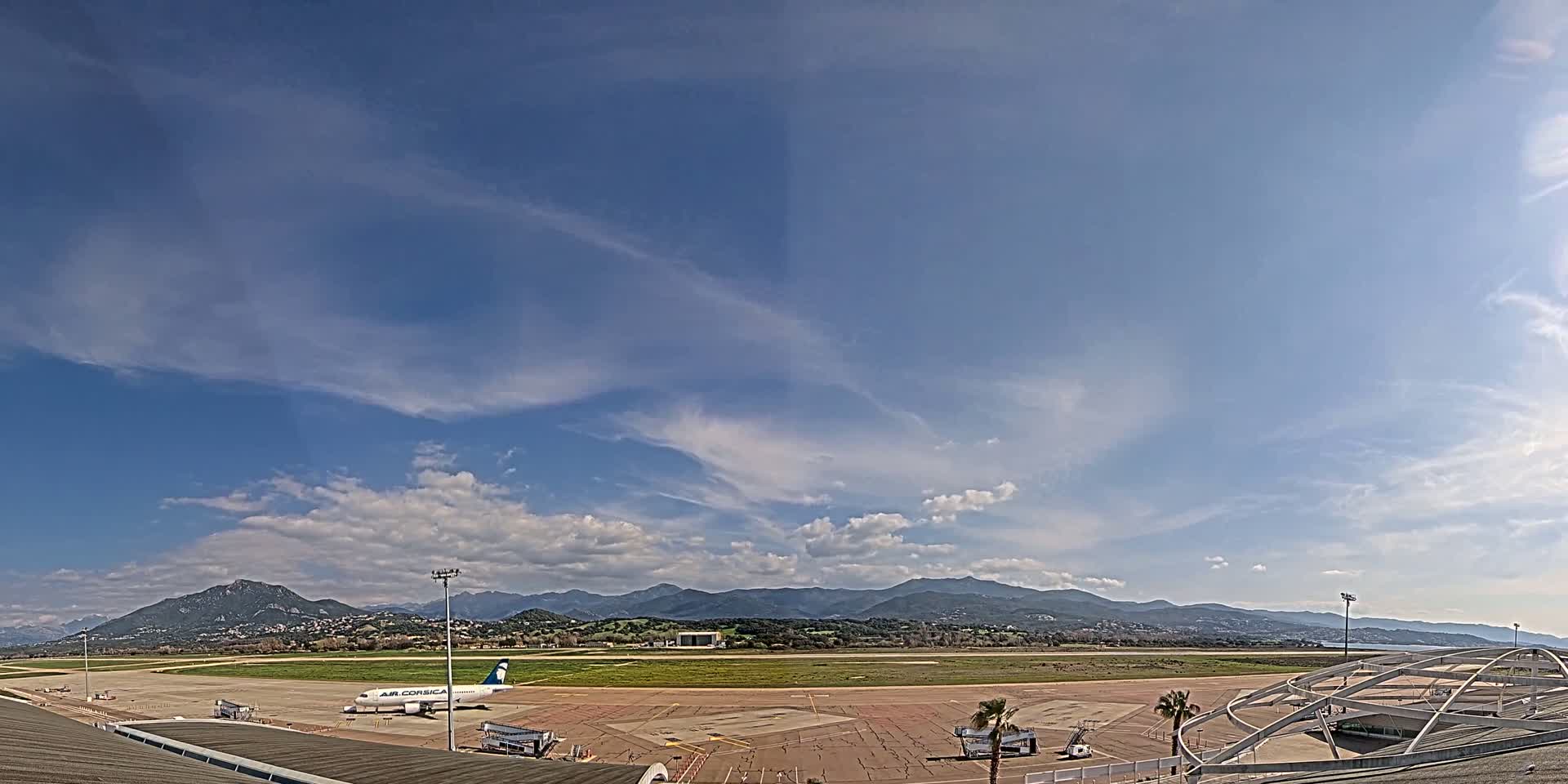 A mostly sunny day with scattered clouds over an airport runway and tarmac, featuring a single airplane and mountains in the distance.