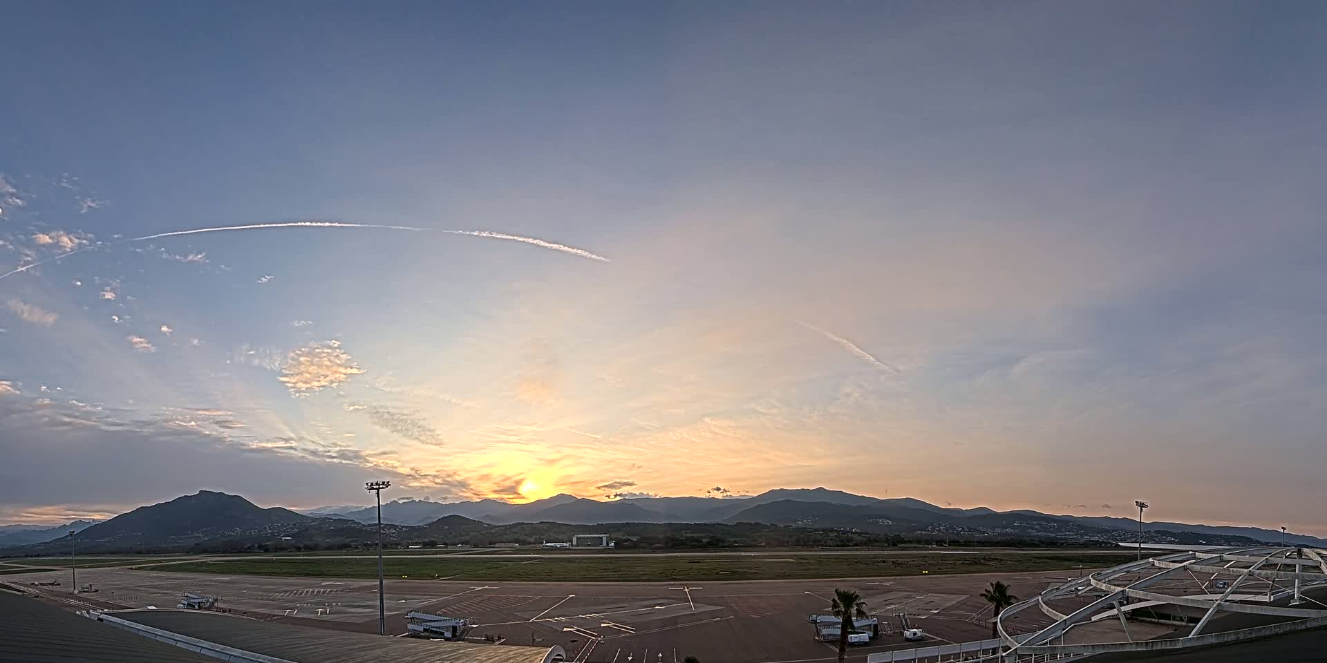 A mostly clear sunset illuminates an airport tarmac and runway, backed by a range of mountains.