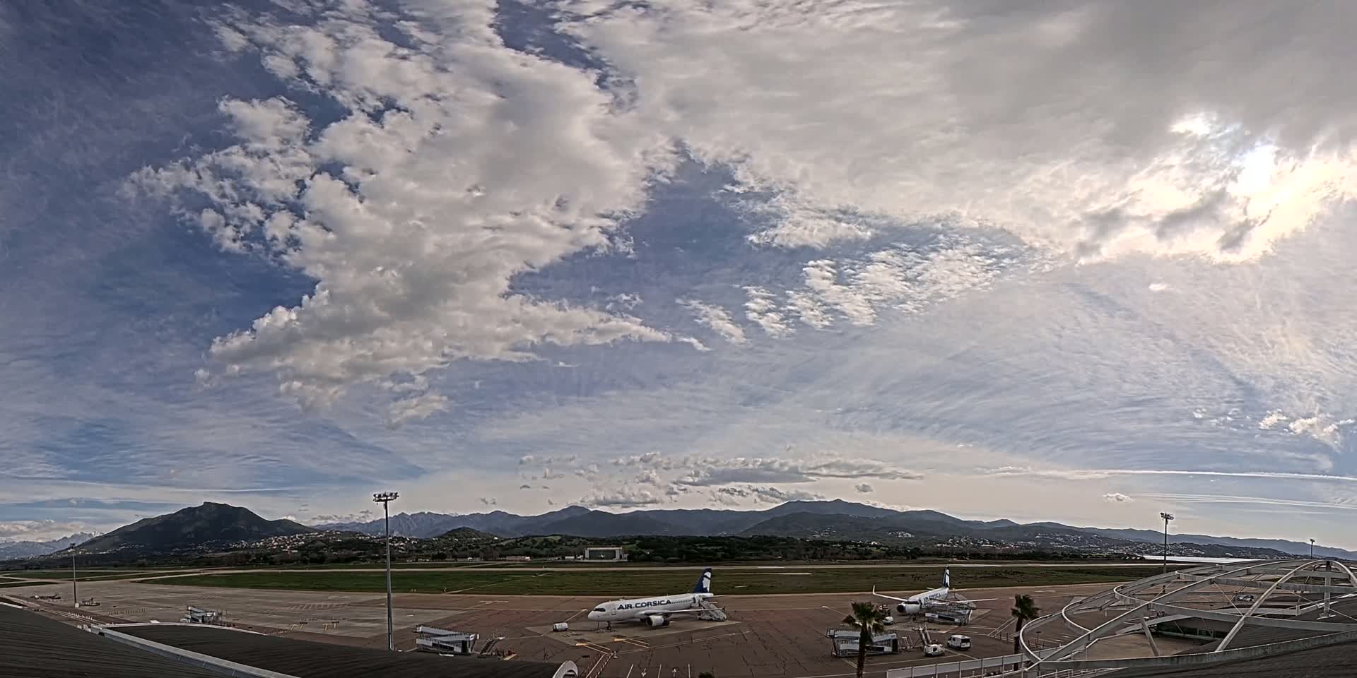 A partly cloudy sky overlooks an airport tarmac with several airplanes parked near airport buildings, and mountains are visible in the distance.