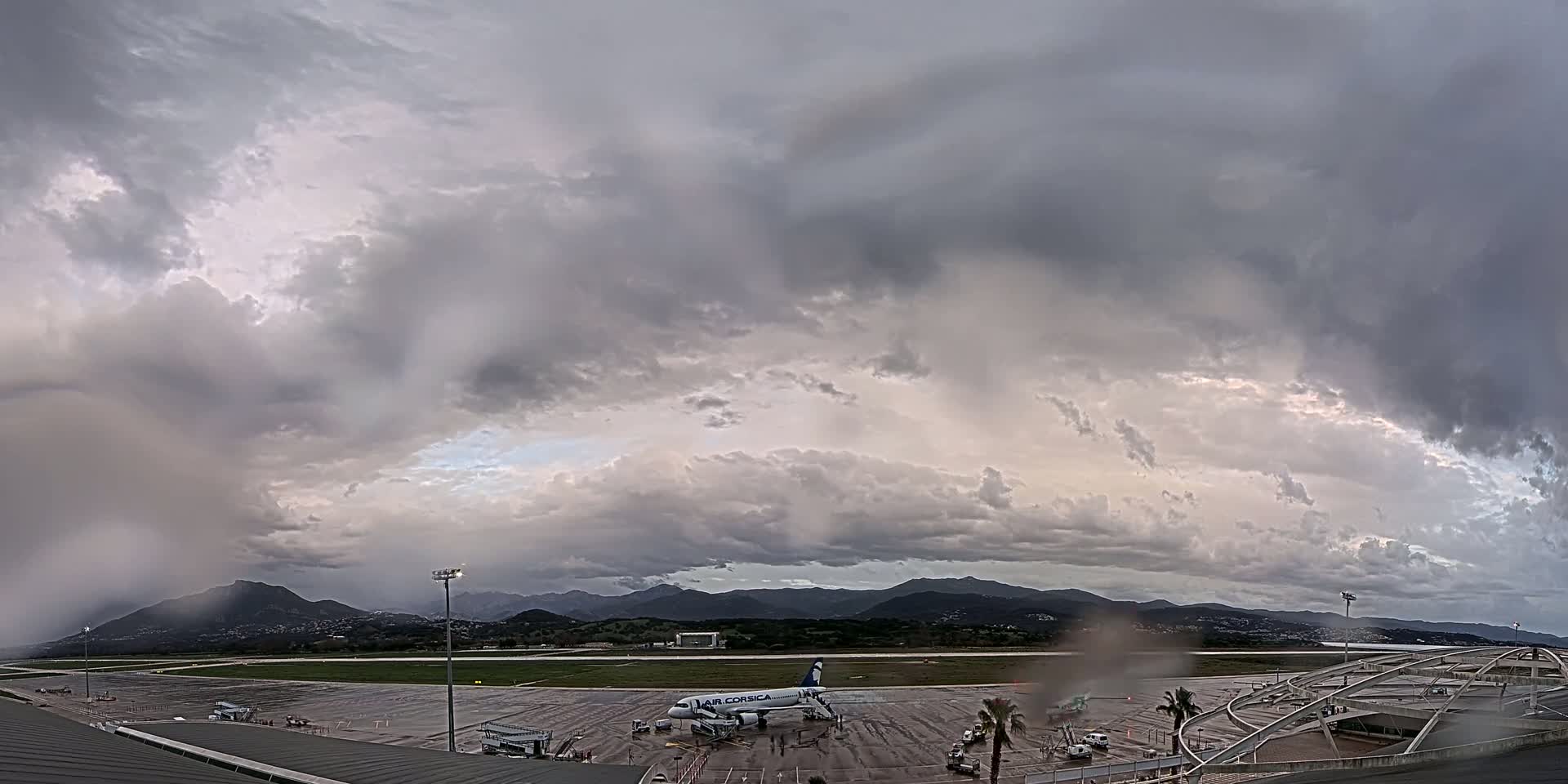 A passenger airplane sits on a wet tarmac at an airport under a stormy, overcast sky, with mountains visible in the distance.
