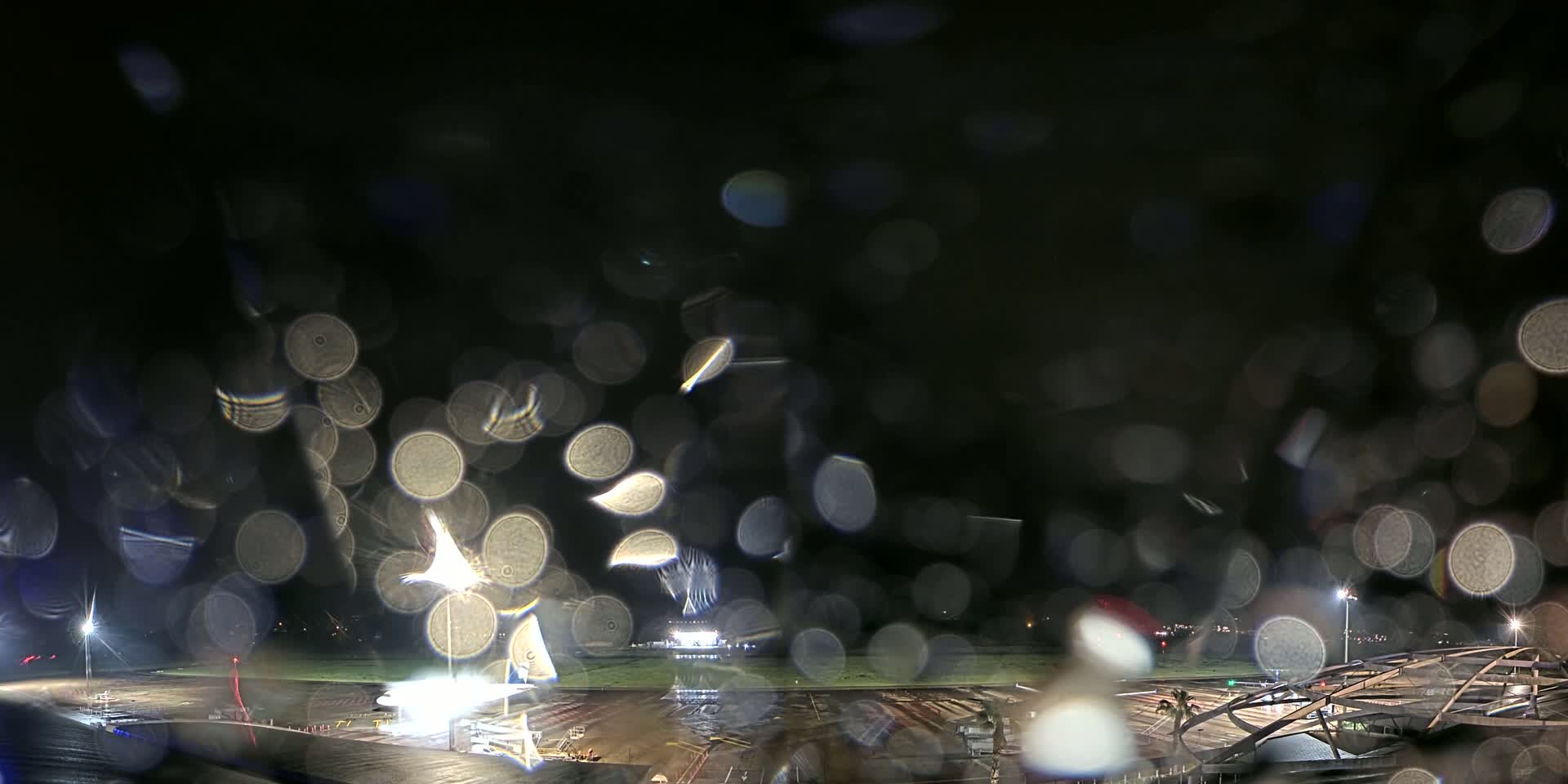 A rain-covered window obscures a nighttime view of a brightly lit airport tarmac and runway.