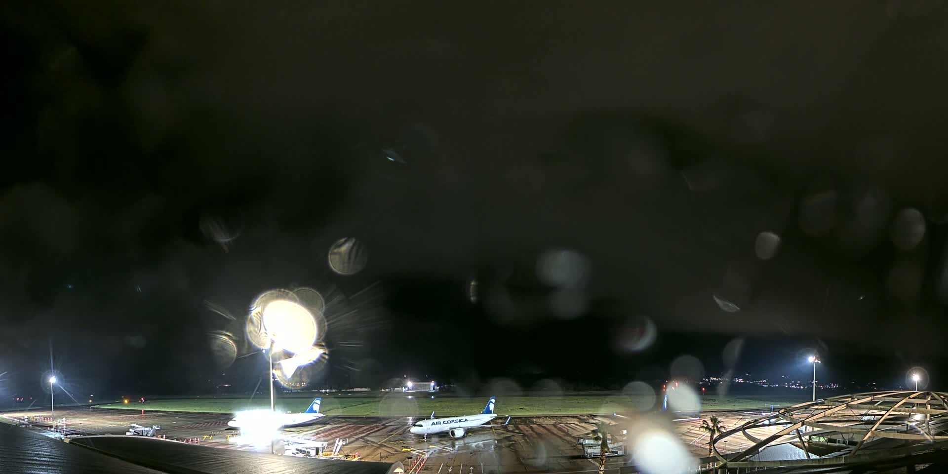 Two airplanes are parked on a rain-soaked airport tarmac at night, viewed through a rain-covered window.