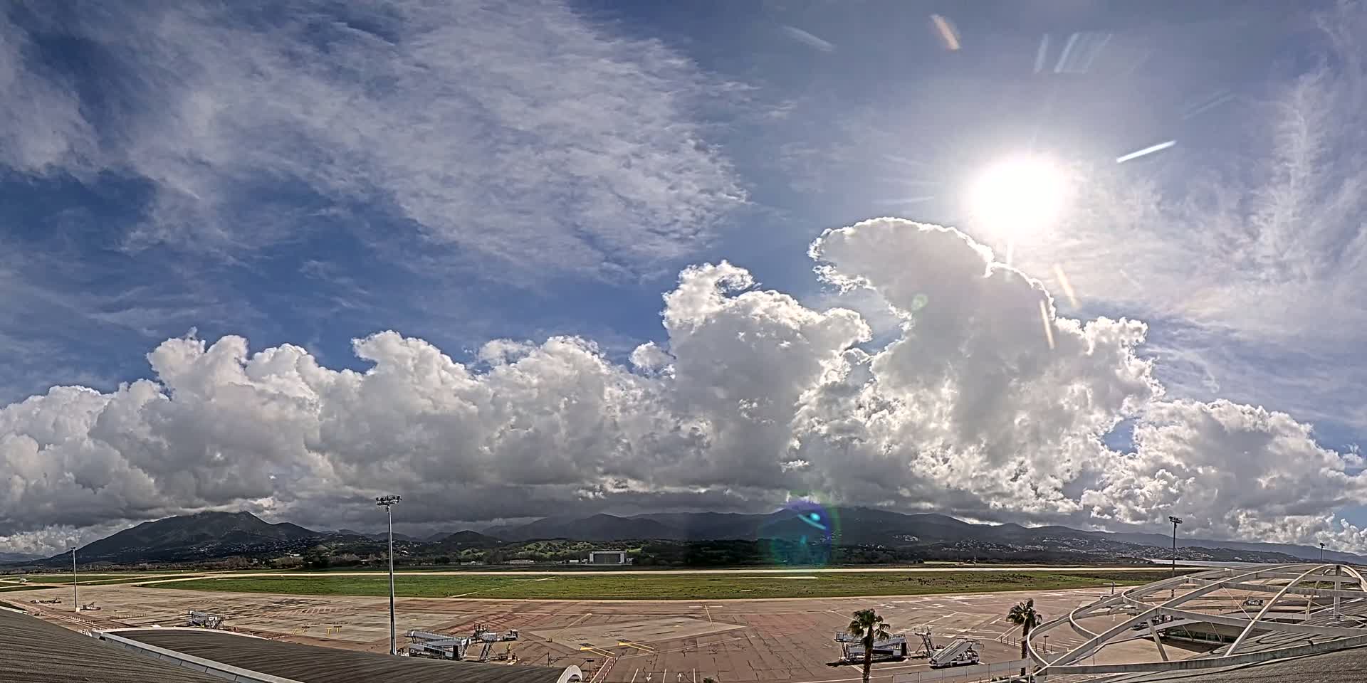 A sunny day with puffy cumulus clouds over an airport tarmac, with mountains visible in the distance.