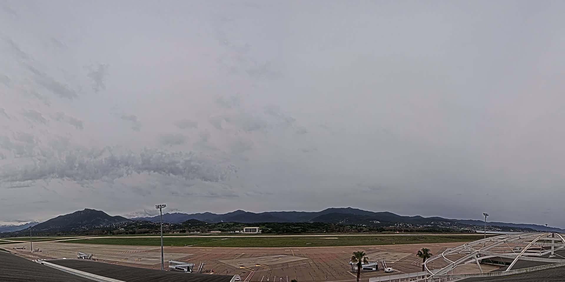 An overcast sky hangs over an airport tarmac and runway, with mountains visible in the distance.