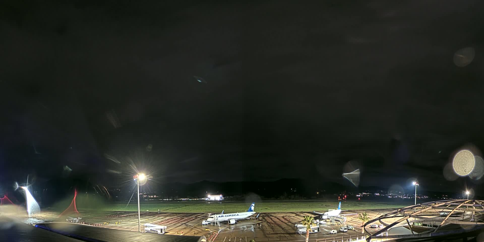A nighttime view of an airport tarmac with a passenger plane and ground support vehicles under a dark, rainy sky.