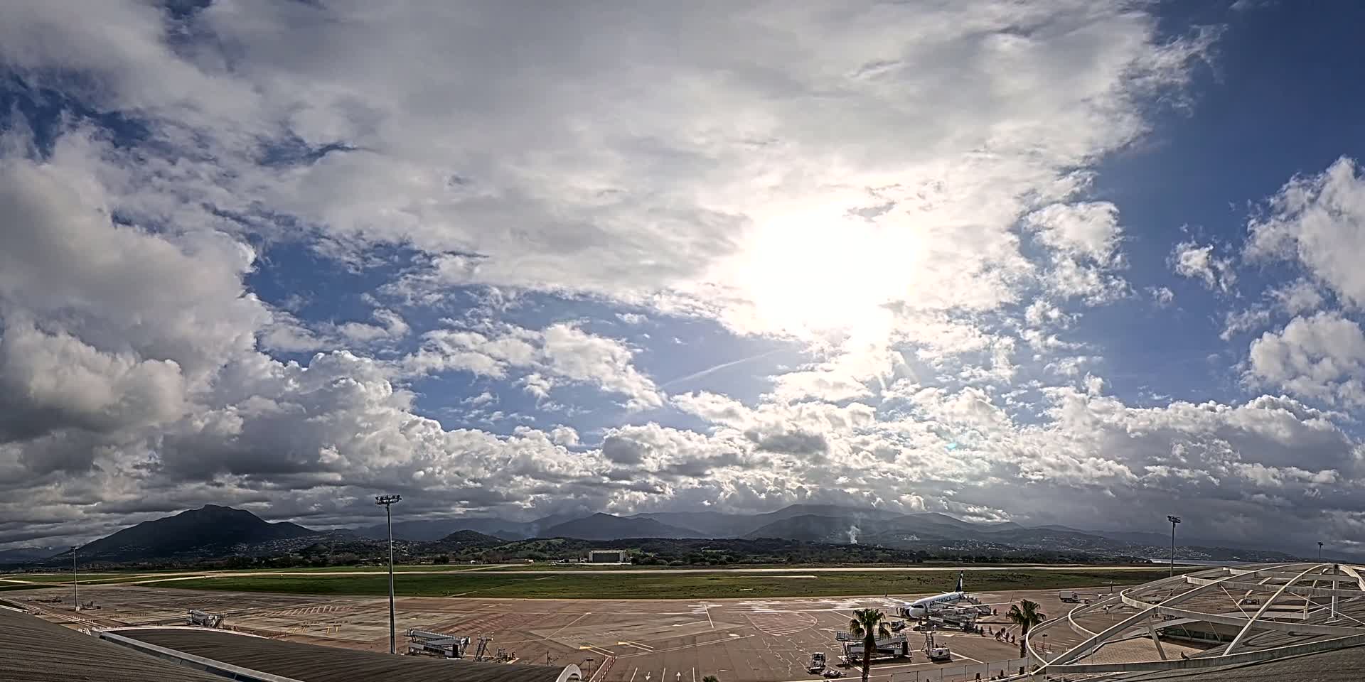 A partly sunny day with puffy clouds over an airport tarmac and runway, backed by distant mountains.
