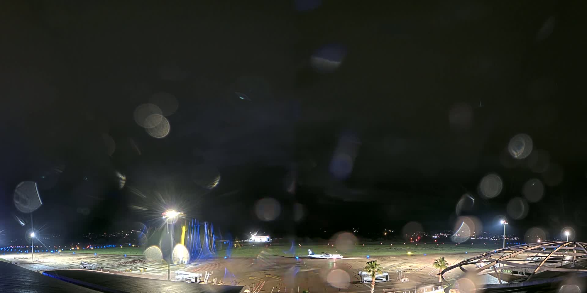 A nighttime view of an airport tarmac with an airplane, viewed through rain-covered glass.