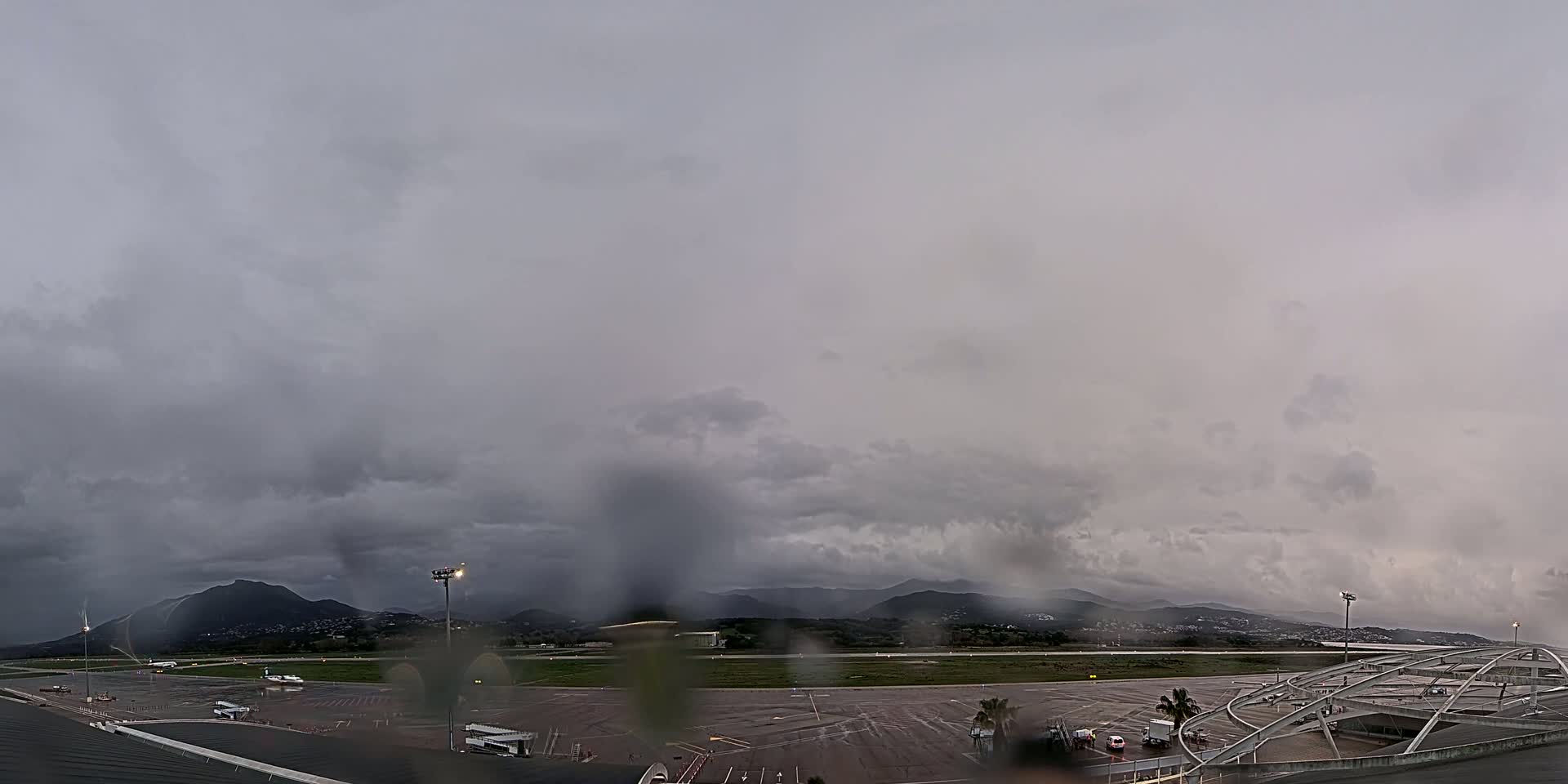 A rain-covered view of an airport tarmac and runway, with dark clouds and mountains in the background.