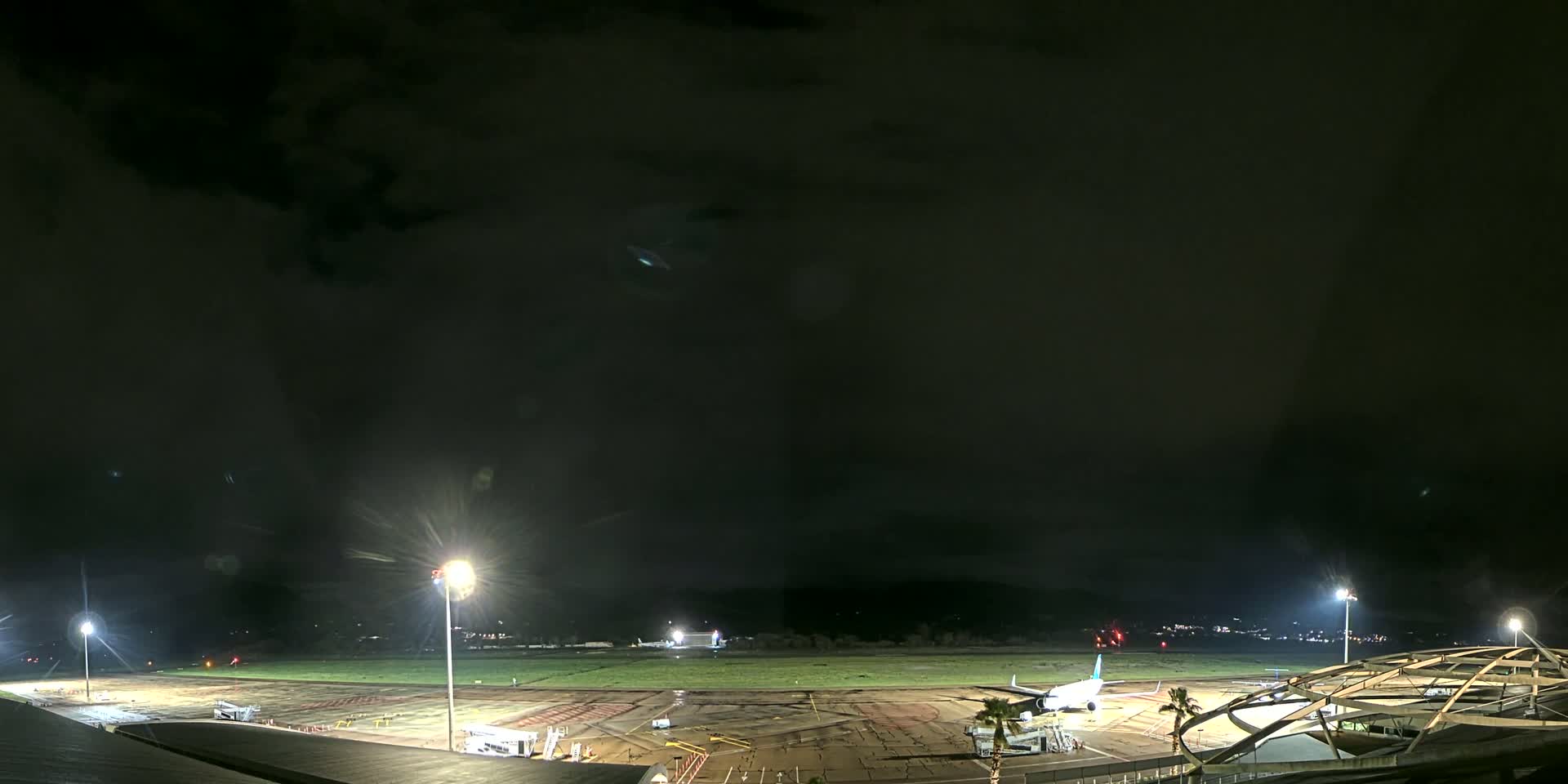 A nighttime view of an airport tarmac with a plane at a gate under a dark, cloudy sky.
