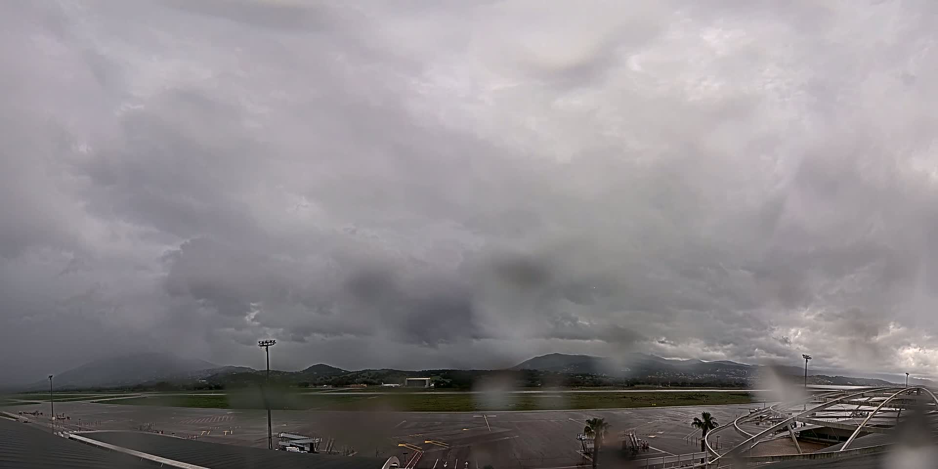 A rain-covered view of an airport tarmac and runway, with dark, stormy clouds overhead and distant mountains visible in the background.