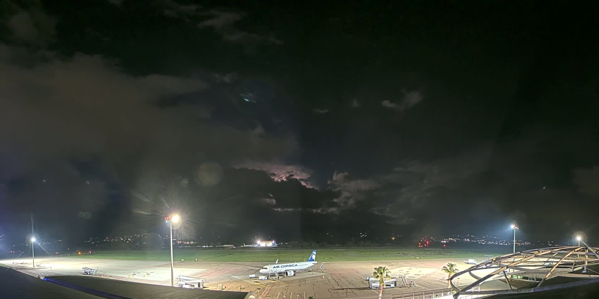 A nighttime view of an airport tarmac with a single airplane parked, under a dark, cloudy sky with hints of lightning.