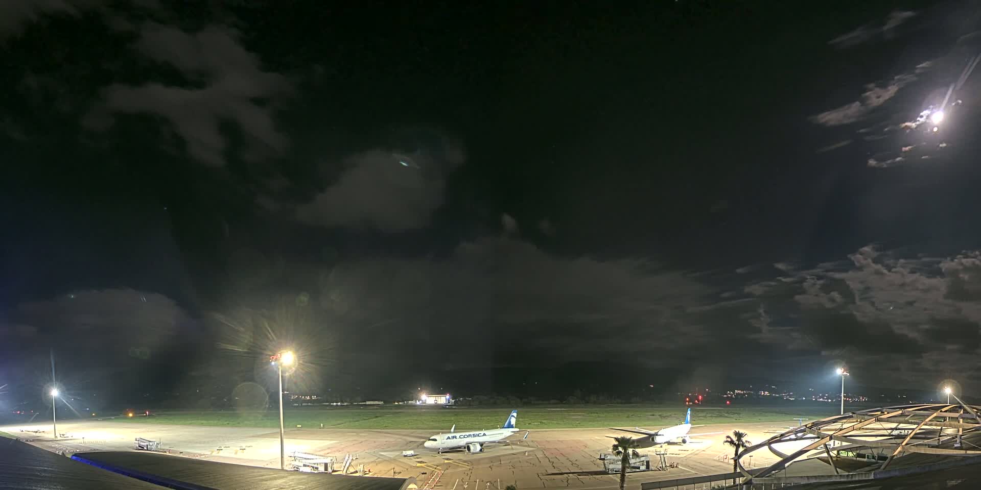 A nighttime view of an airport tarmac with two airplanes parked, illuminated by bright lights under a partly cloudy sky.