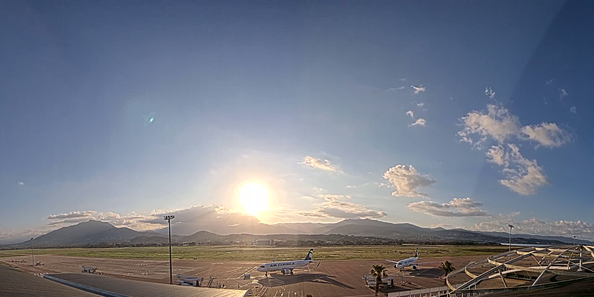 Two airplanes sit on a tarmac at sunset under a partly cloudy sky with mountains in the distance.