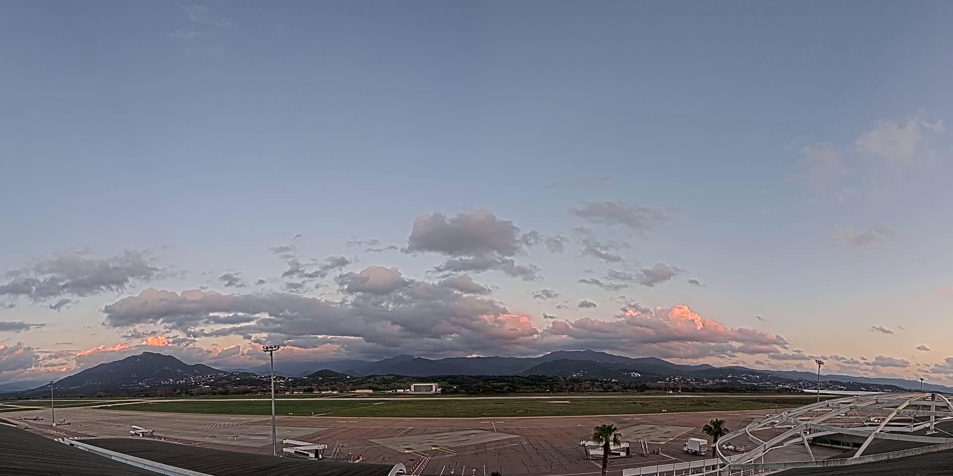 An airport tarmac with a runway and surrounding mountains is visible under a partly cloudy sky at sunset.