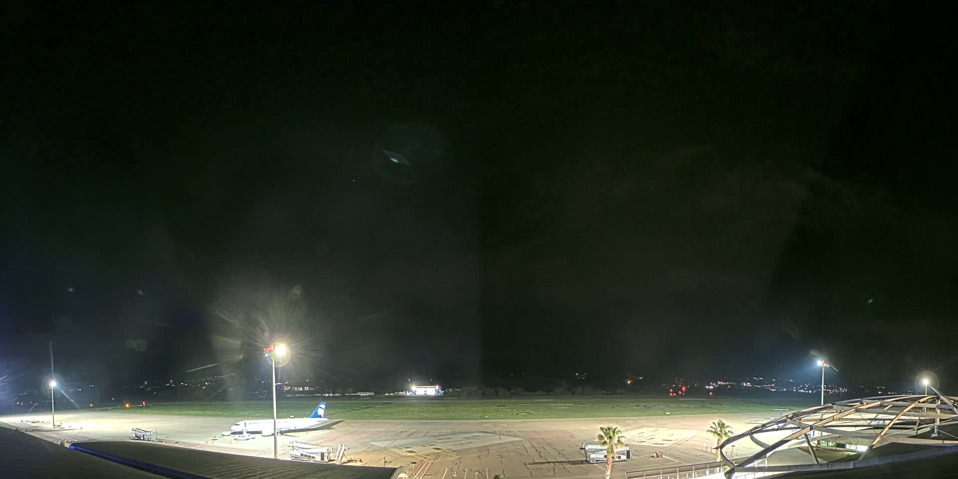 A nighttime view of an airport tarmac with a plane parked near a terminal building under a mostly clear, dark sky.