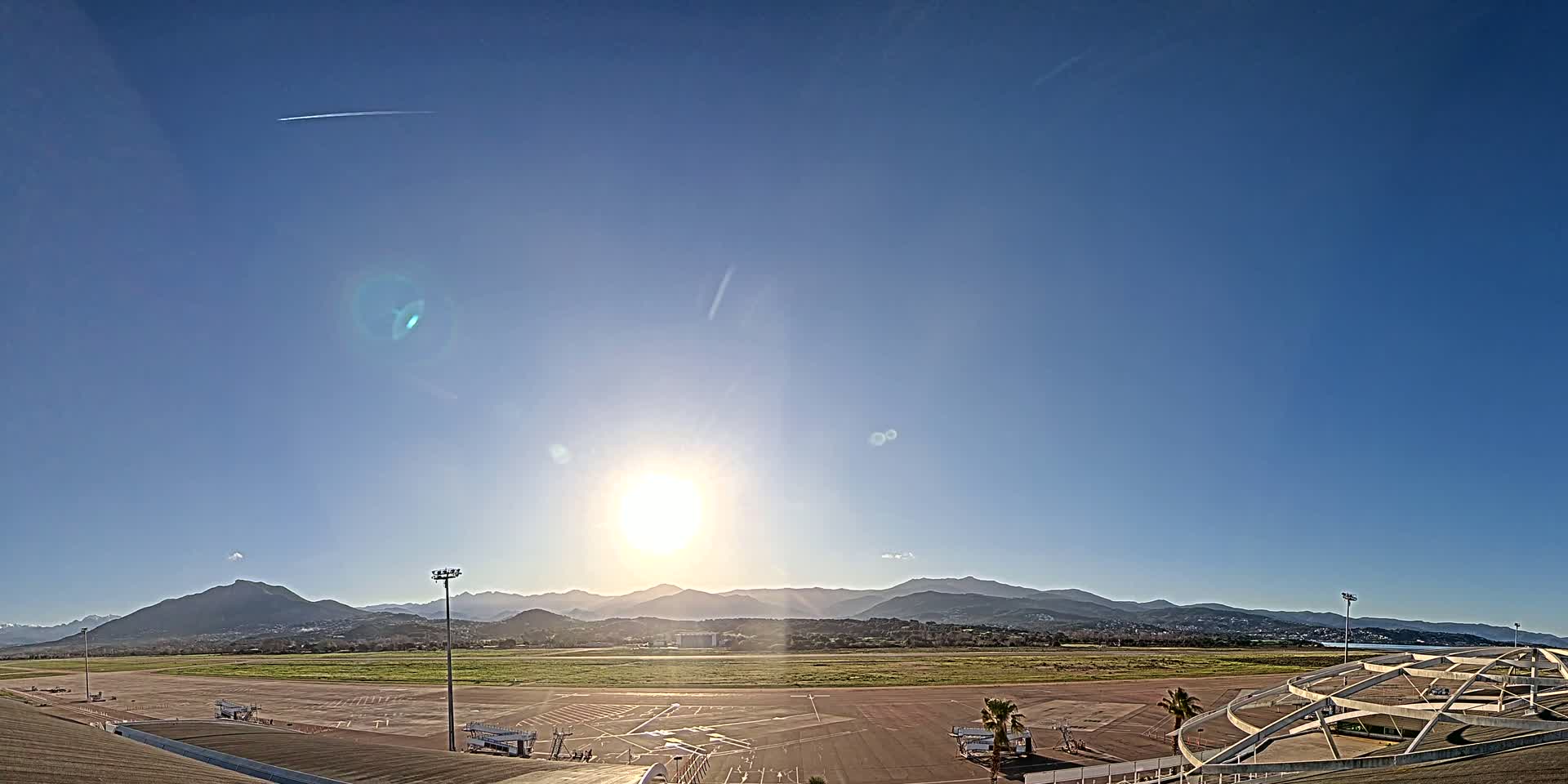 Under a clear, bright sunny sky, an airport tarmac with several aircraft parked near jet bridges is situated in front of a range of distant mountains.