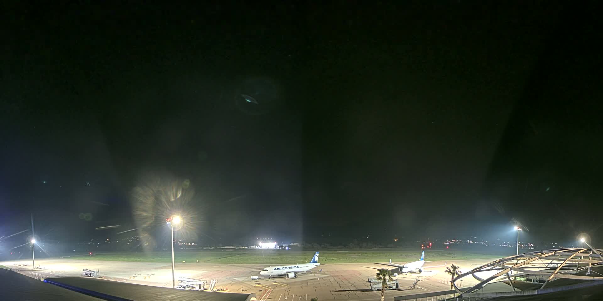 At night, an airport tarmac is illuminated, showing two airplanes parked near airport buildings under a clear, dark sky.