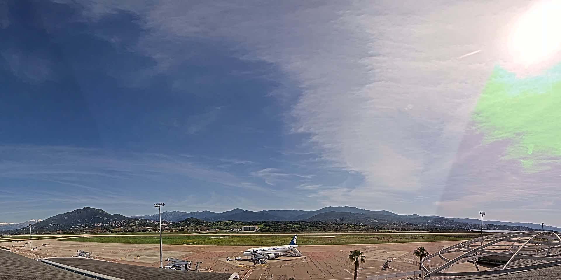 A partly cloudy sky overlooks an airport tarmac with a single airplane parked at a gate, backed by a range of mountains in the distance.