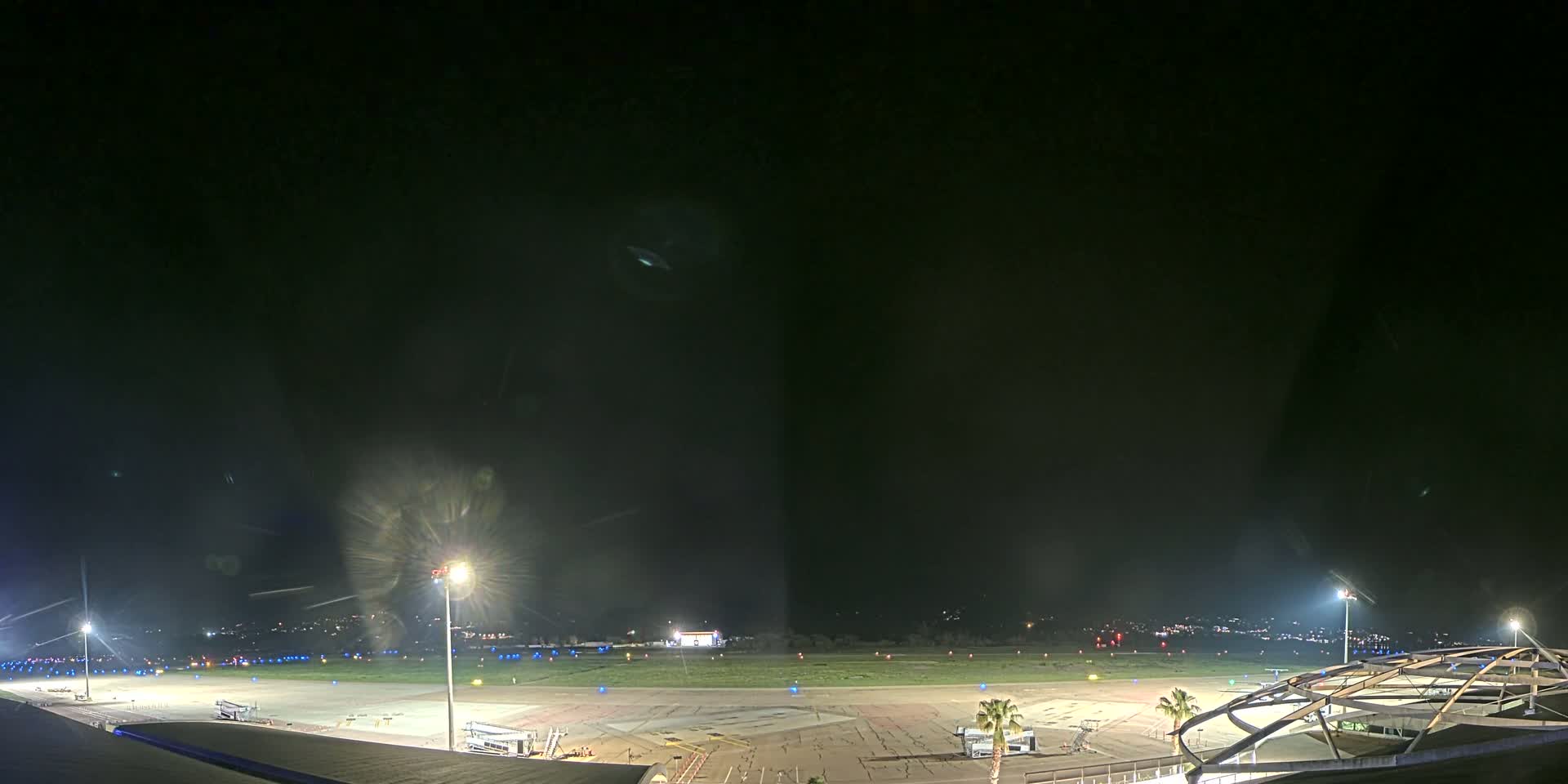 A nighttime view of an airport tarmac, illuminated by floodlights, with a portion of a terminal building visible in the foreground under a clear, dark sky.