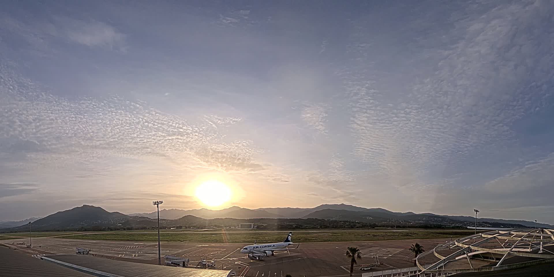 A passenger airplane sits on an airport tarmac at sunrise under a mostly clear sky with scattered clouds, mountains visible in the distance.