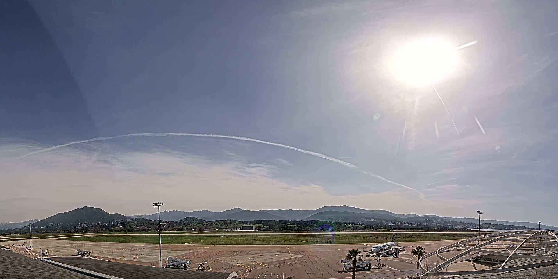 A sunny day at an airport shows a tarmac with several airplanes, jet bridges, and a distant mountain range under a mostly clear sky with contrails.