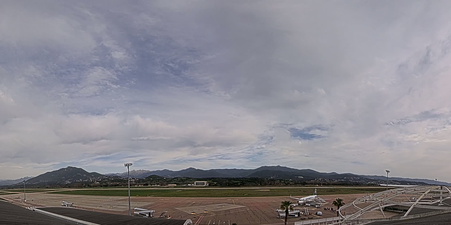 An airport tarmac with a large airplane and several buildings is visible under a mostly cloudy sky, with mountains in the distance.