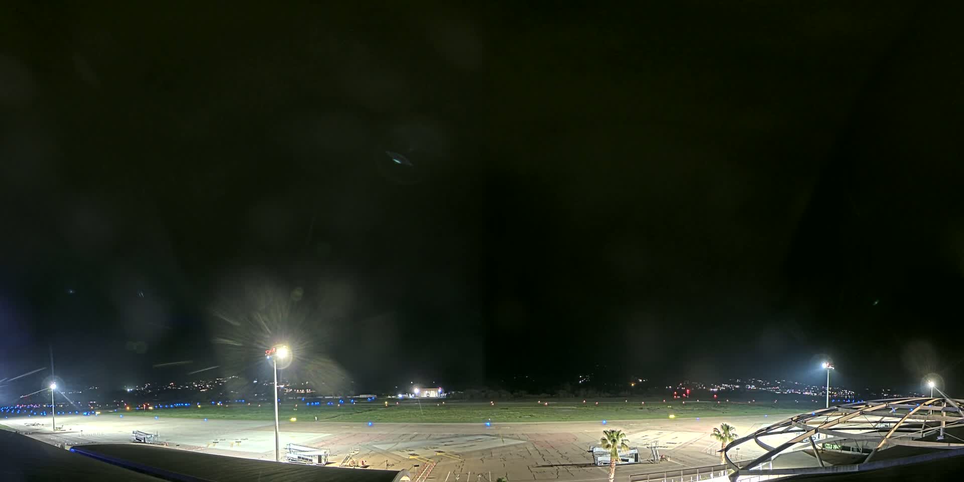 A nighttime view of an airport tarmac with several illuminated areas, some distant lights, and a partially visible structure at the bottom right.