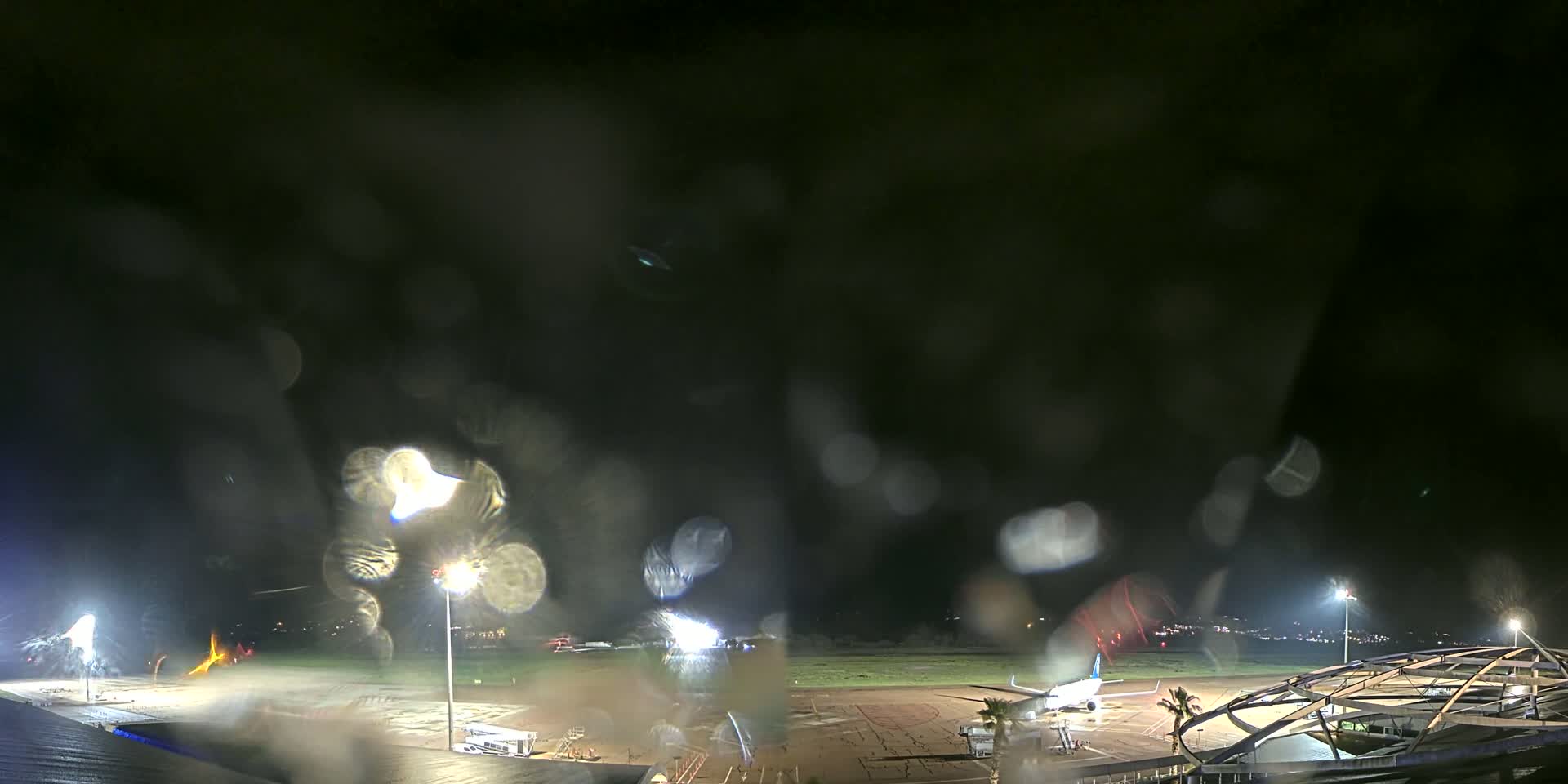 A nighttime view of an airport tarmac with a plane parked near a terminal building, seen through rain-covered glass.