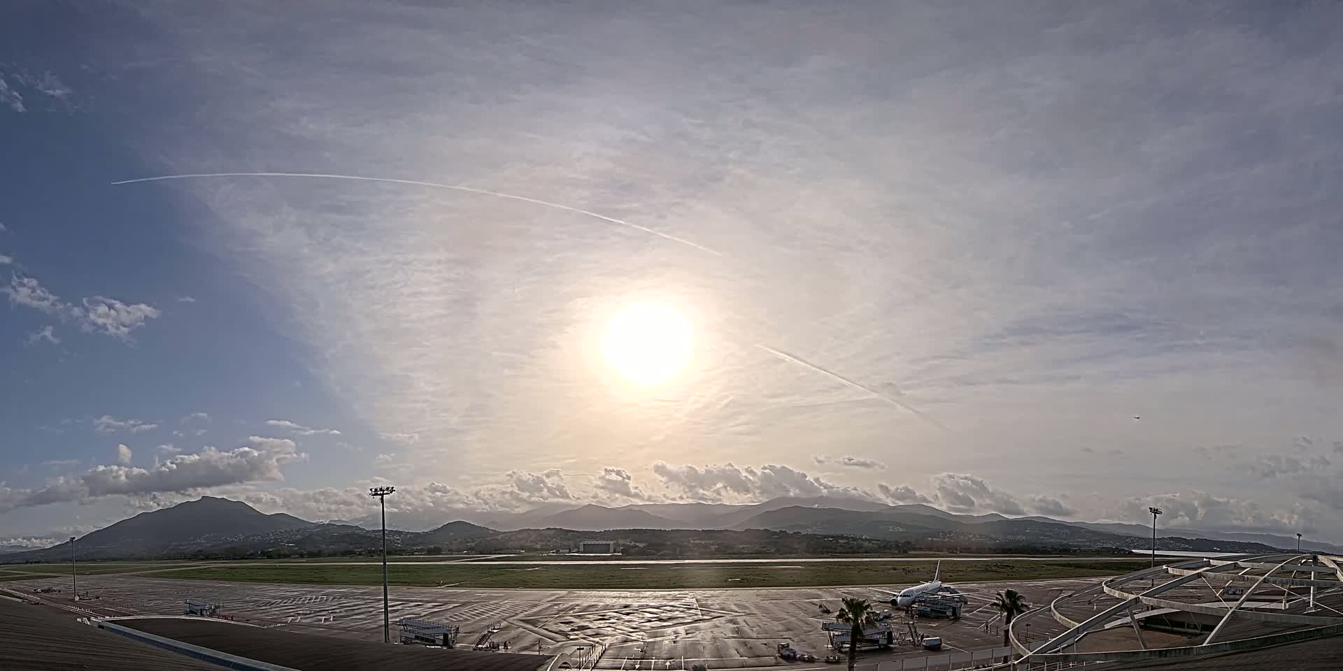A partly cloudy day reveals a sunlit airport tarmac with a plane parked at a gate, backed by mountains in the distance.