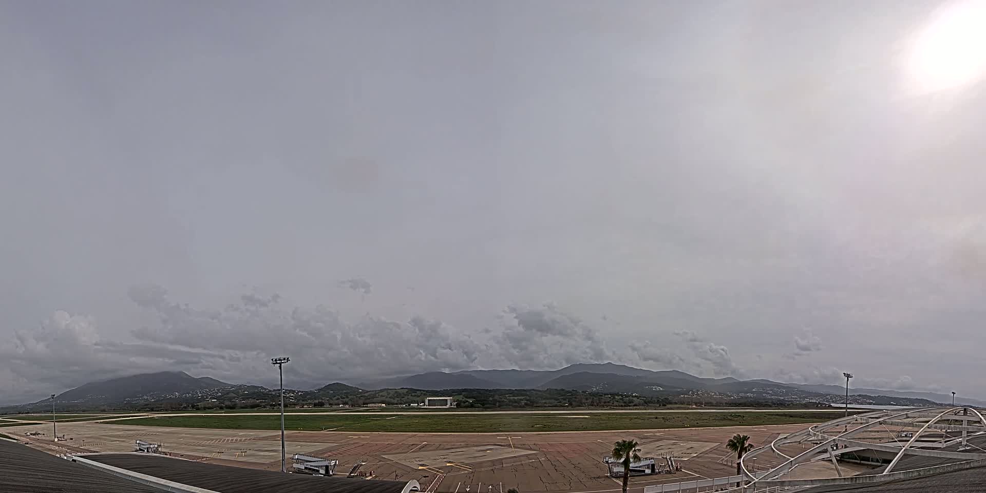 An airport tarmac and runway are visible under an overcast sky, with mountains in the background.