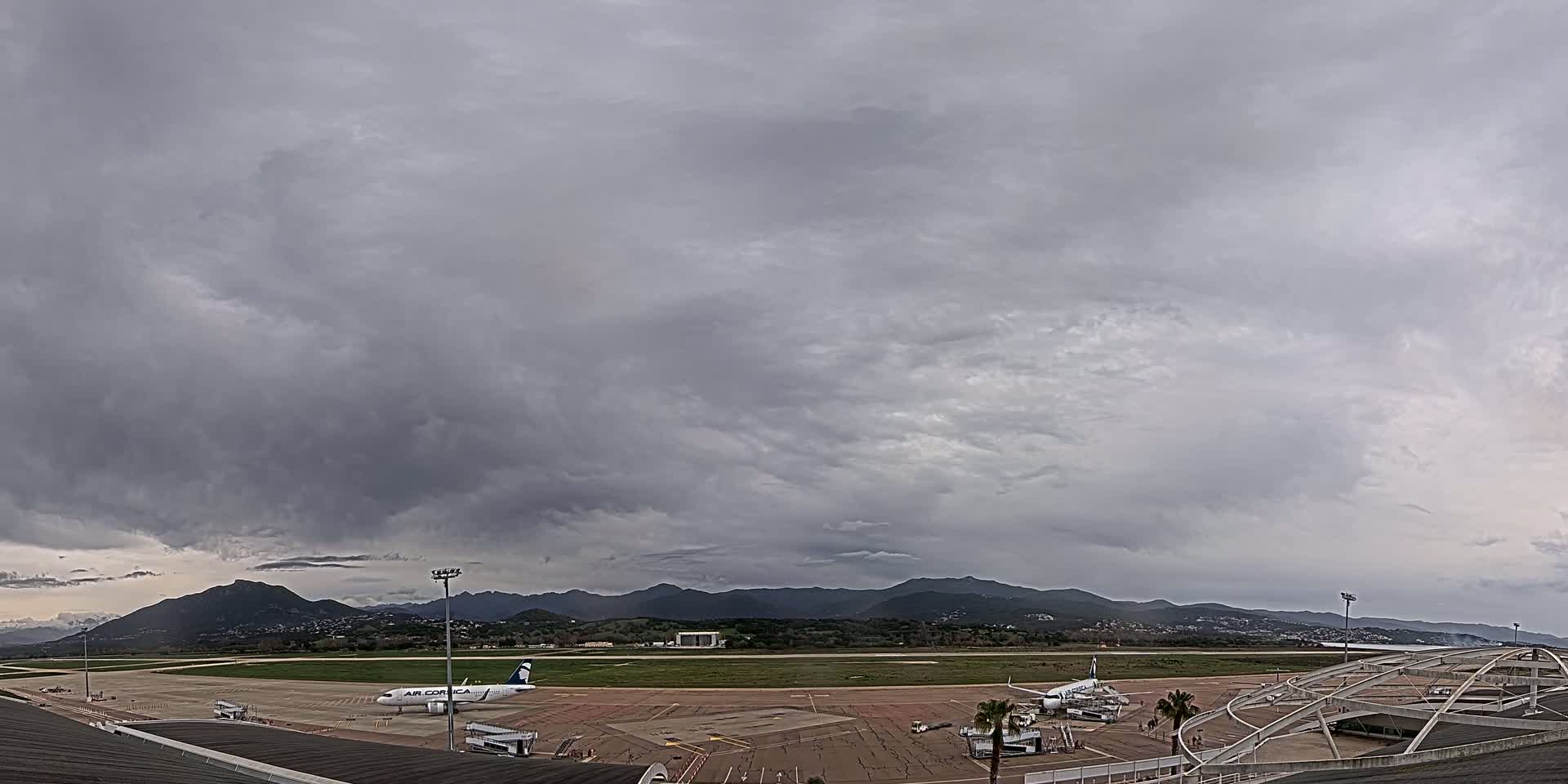 A mostly cloudy sky hangs over an airport tarmac where a plane is parked, with mountains visible in the distance.