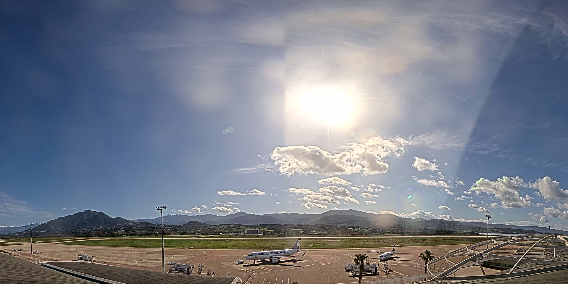 A sunny day at an airport shows two airplanes parked on the tarmac with mountains in the background.