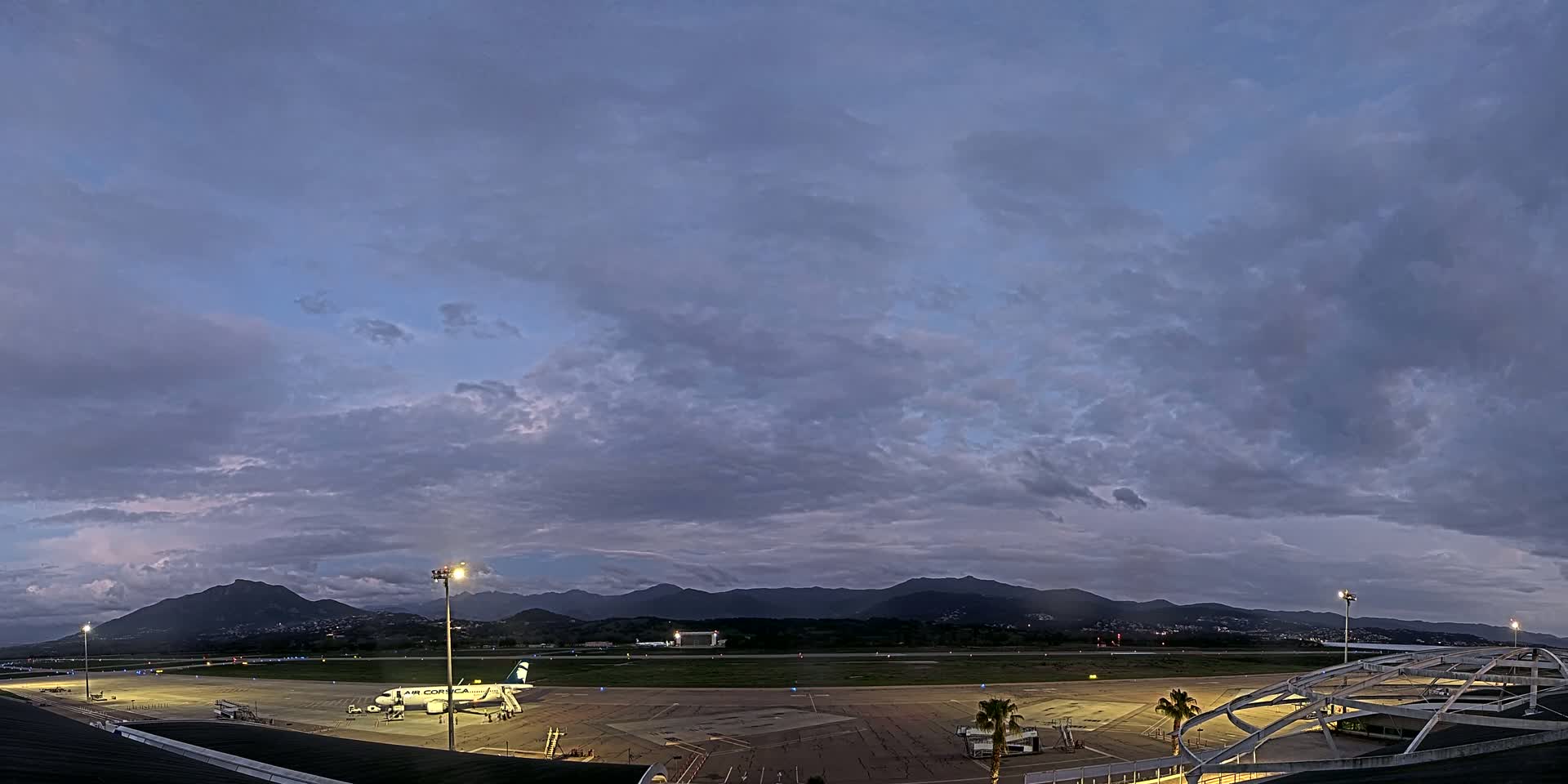 An airplane sits on an airport tarmac at dusk under a cloudy sky, with mountains visible in the distance.