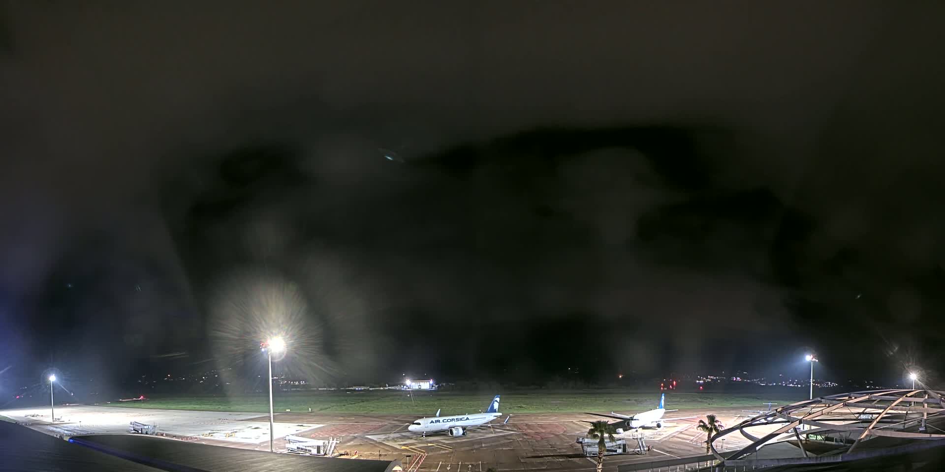 A nighttime view of an airport tarmac under a dark, cloudy sky, showing two airplanes parked near terminal buildings and illuminated by airport lights.