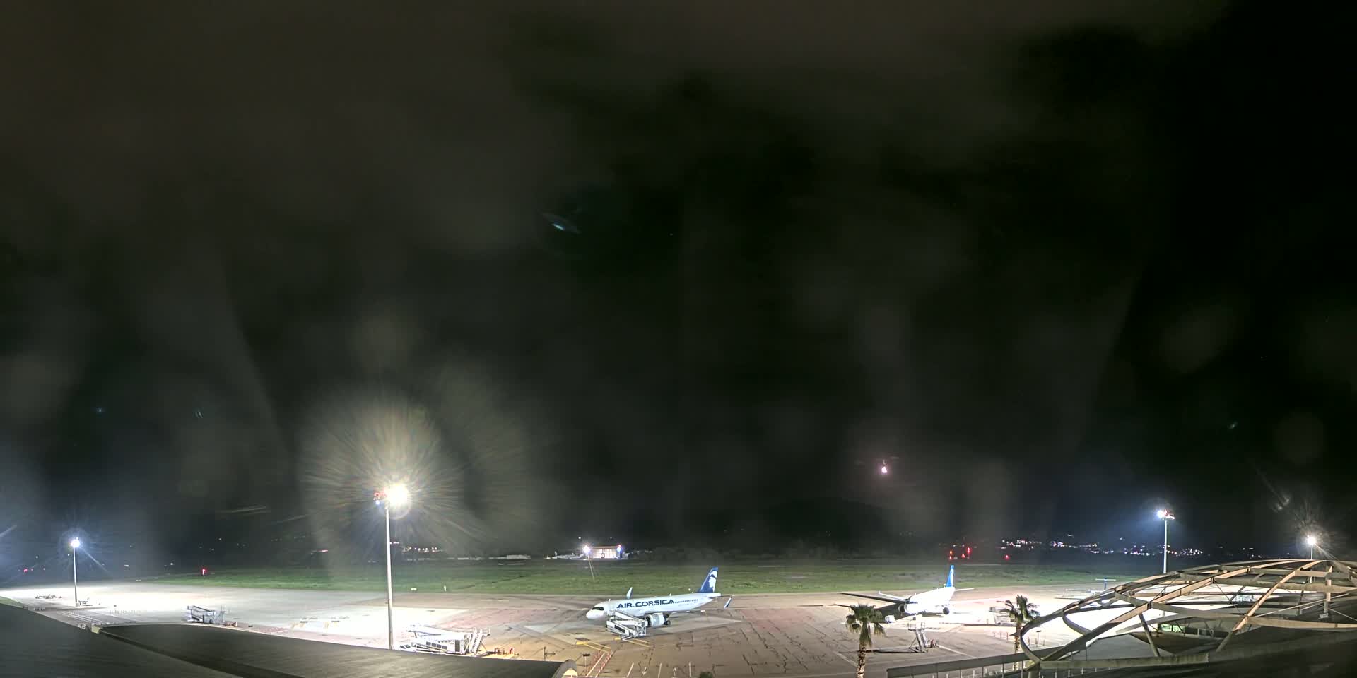 A nighttime view of an airport tarmac with two airplanes parked, under a dark, cloudy sky with light rain or mist.