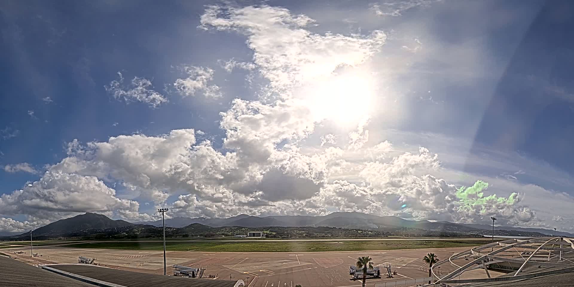 A sunny day with puffy cumulus clouds over an airport tarmac, with mountains visible in the distance.
