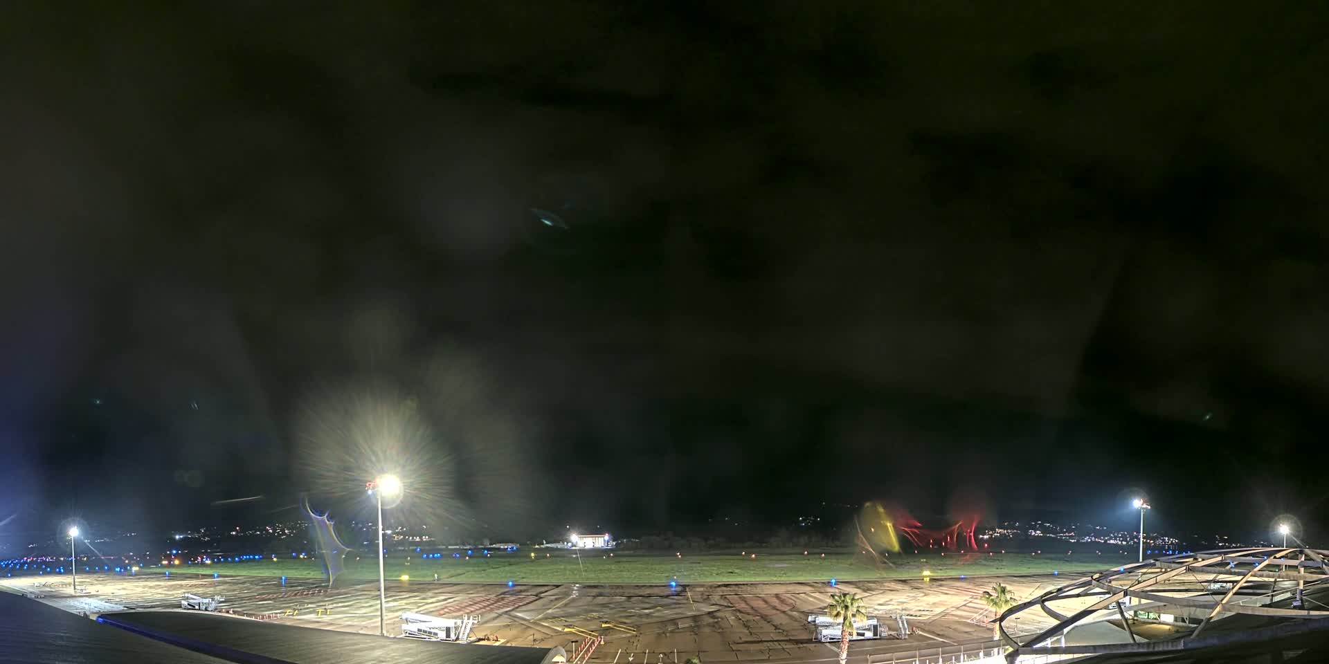 A nighttime view of an airport tarmac under a dark, cloudy sky, illuminated by bright lights.