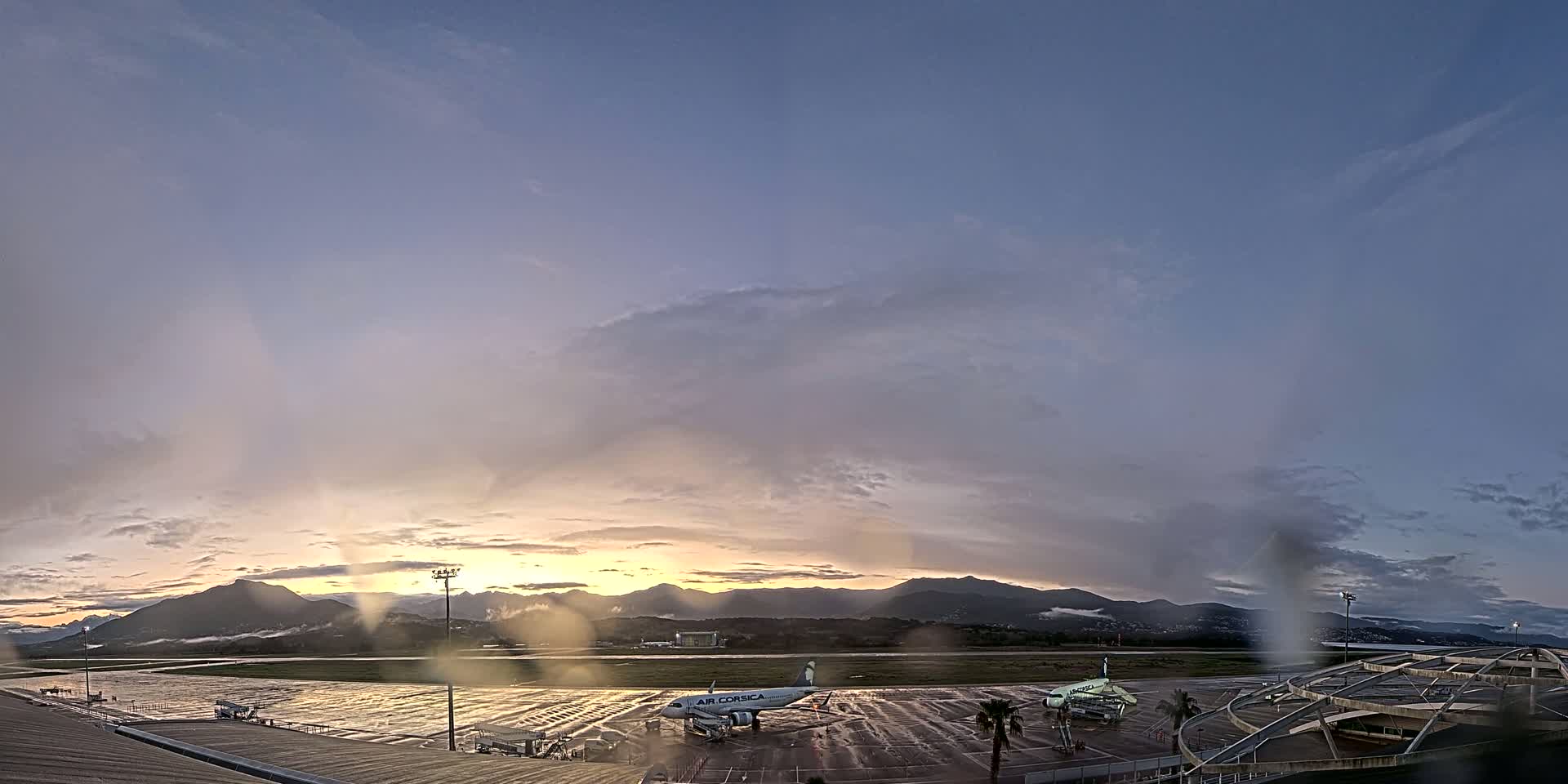 A wet airplane sits on an airport tarmac at sunrise, with rain-streaked windows obscuring a view of mountains in the distance.