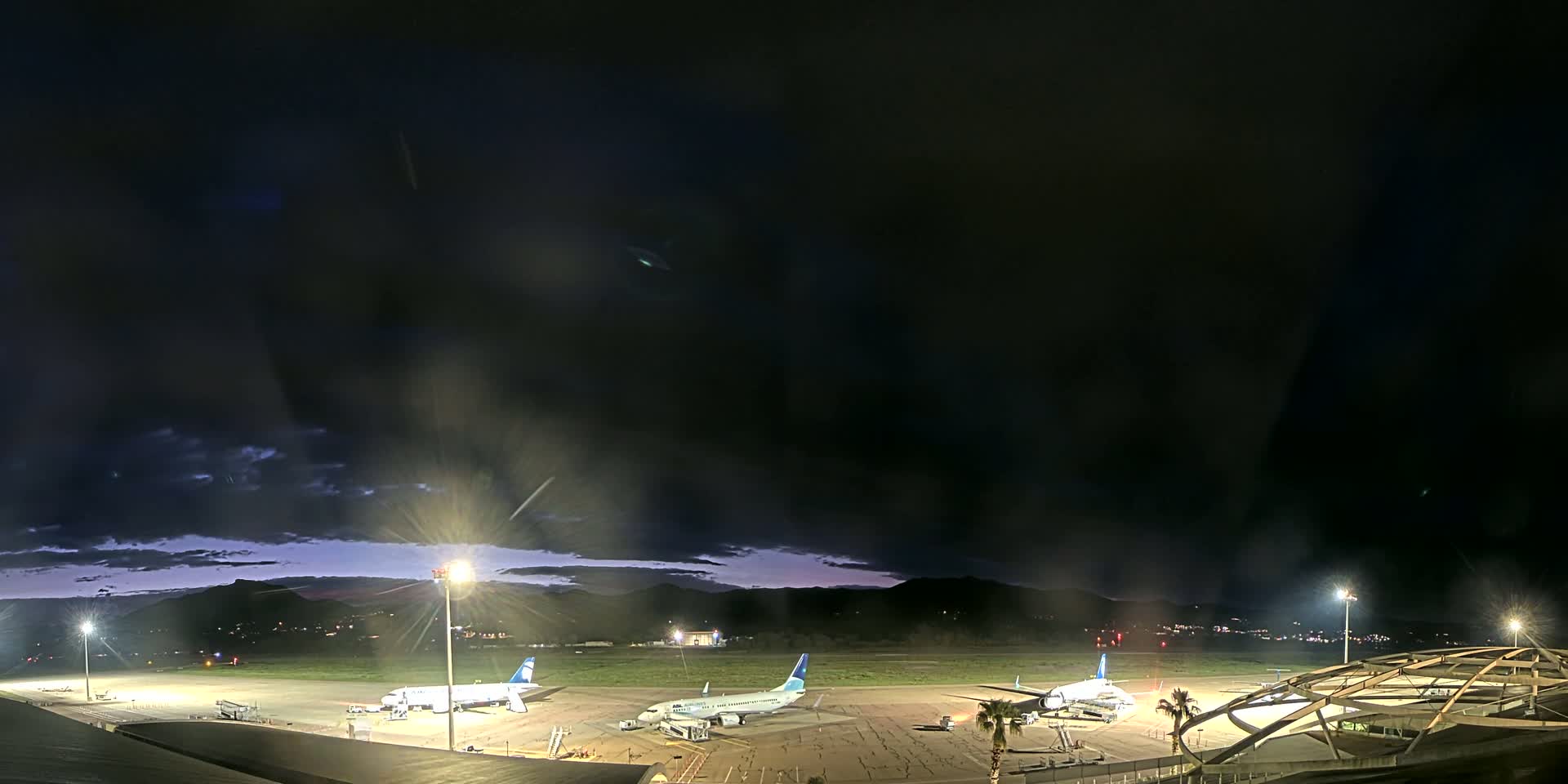 Three airplanes are parked on a dark airport tarmac at night under a mostly cloudy sky.