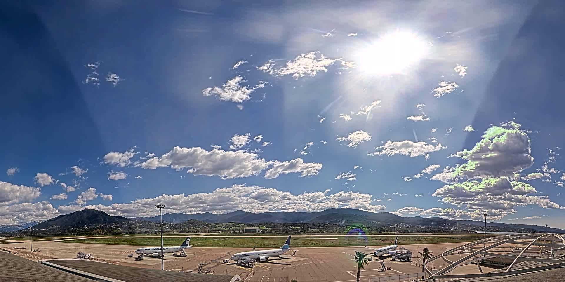 Three airplanes are parked on a tarmac at an airport under a bright, sunny sky with scattered cumulus clouds and mountains in the background.