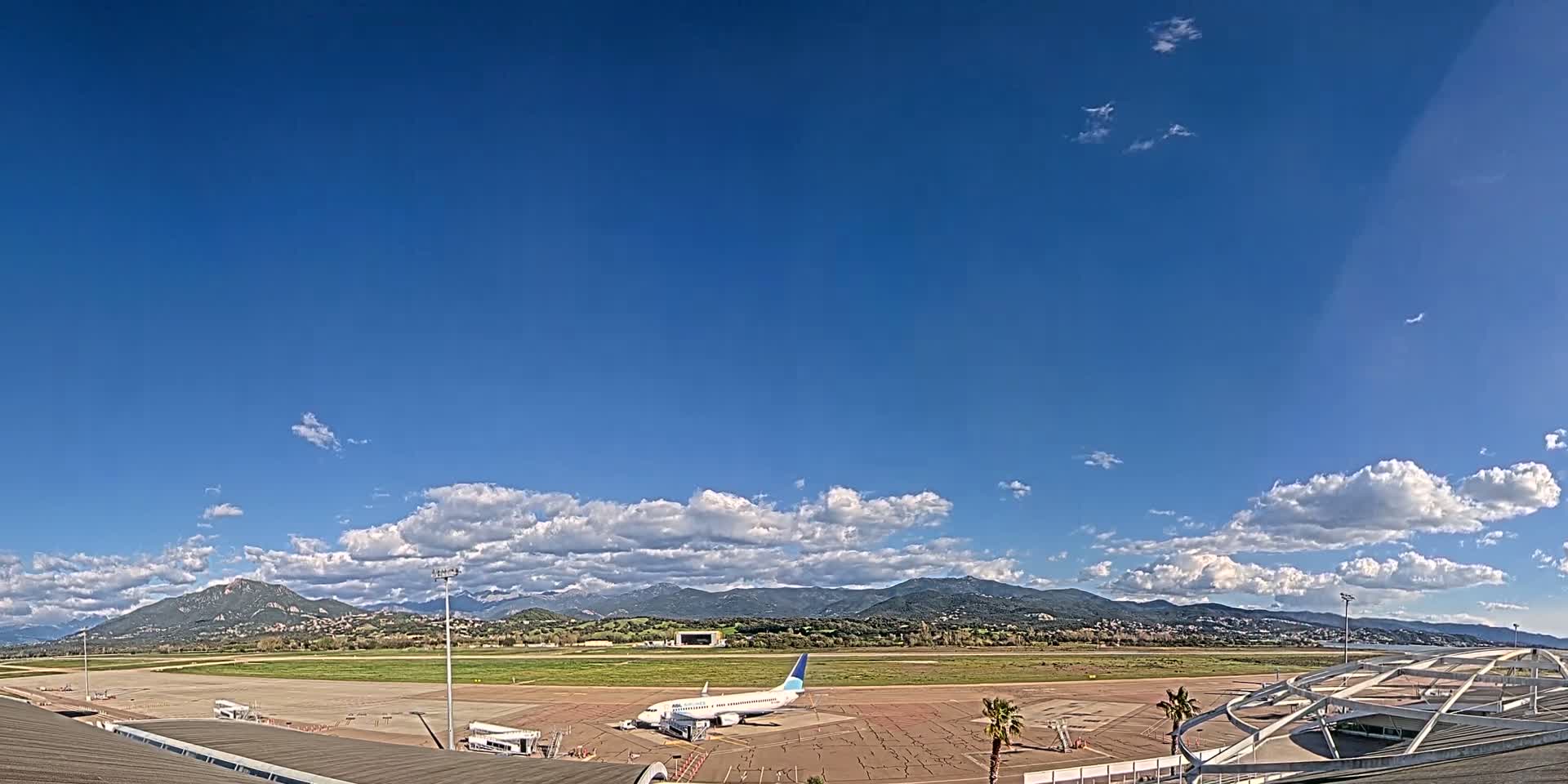 A passenger airplane sits on a tarmac at an airport under a mostly sunny sky with scattered clouds and mountains in the distance.