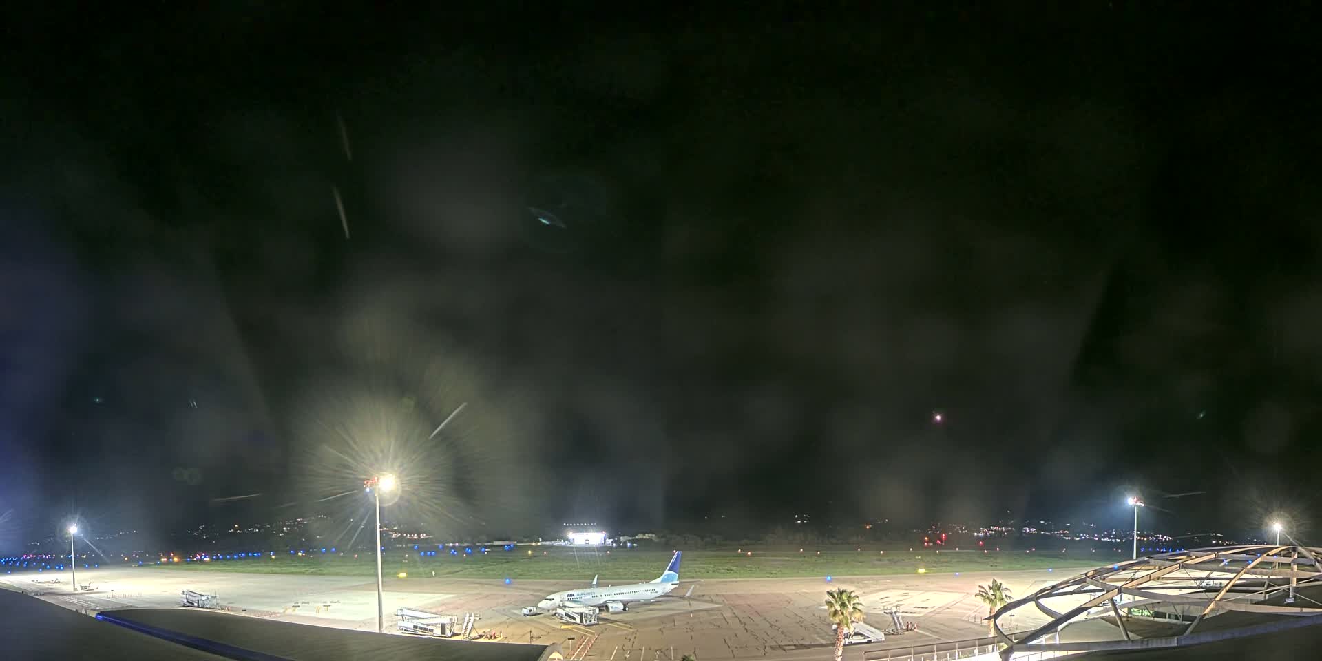 A nighttime view of an airport tarmac with a single airplane parked at a gate, under a dark, hazy sky.