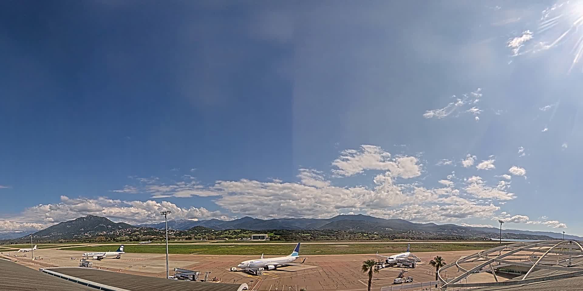 Several airplanes are parked on a tarmac at an airport under a mostly sunny sky with scattered clouds and mountains in the distance.