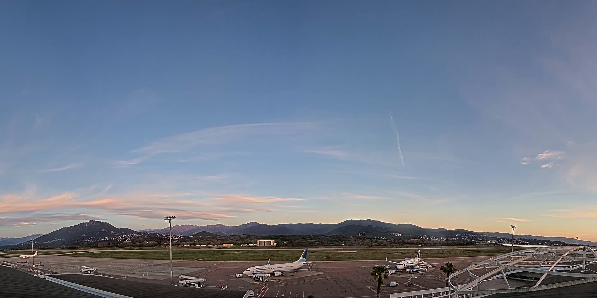 An airplane sits on a tarmac at an airport under a mostly clear sky at sunset, with mountains visible in the distance.