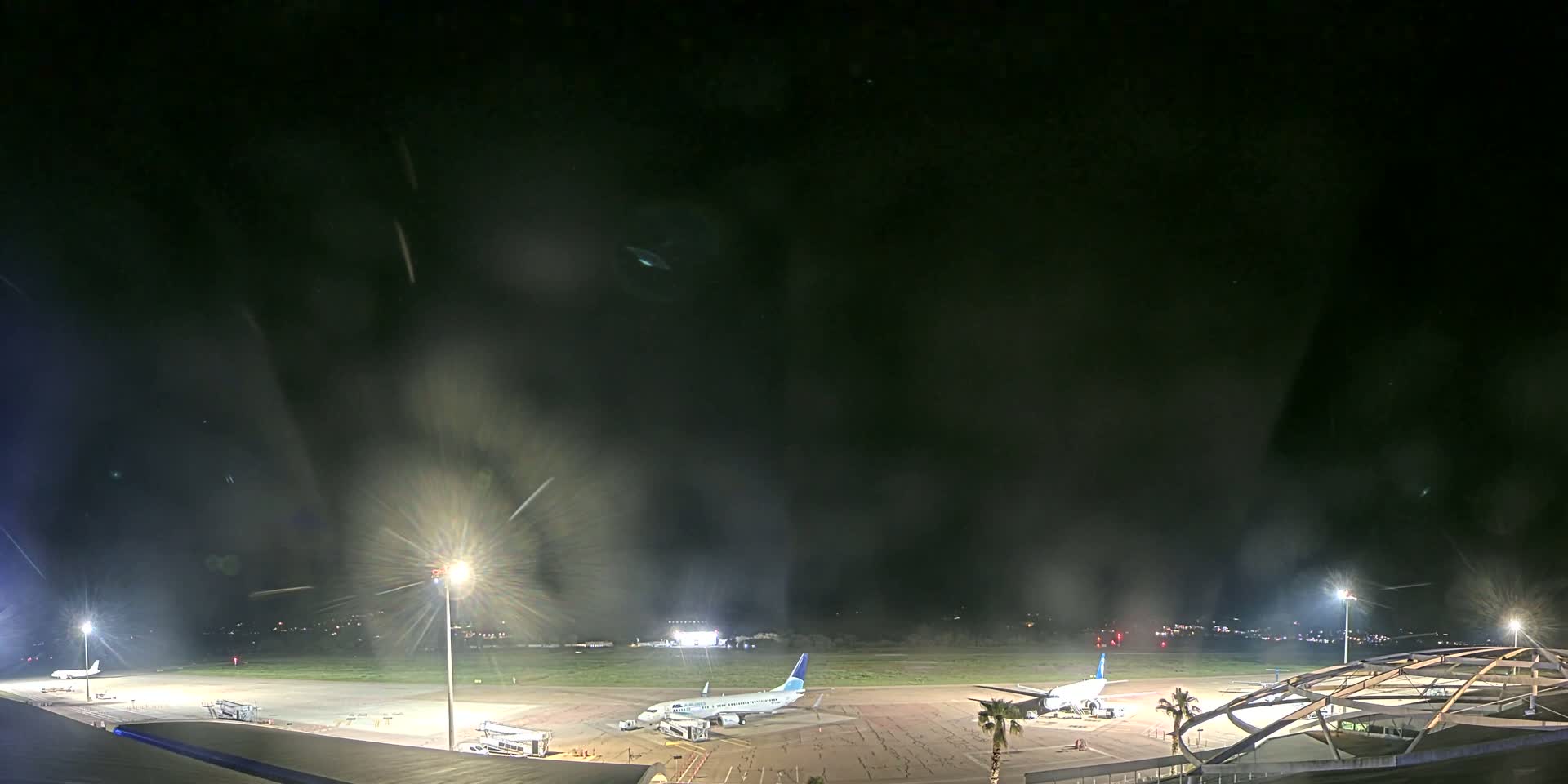 At night, under a dark, possibly hazy sky, several airplanes are parked on a brightly lit airport tarmac.