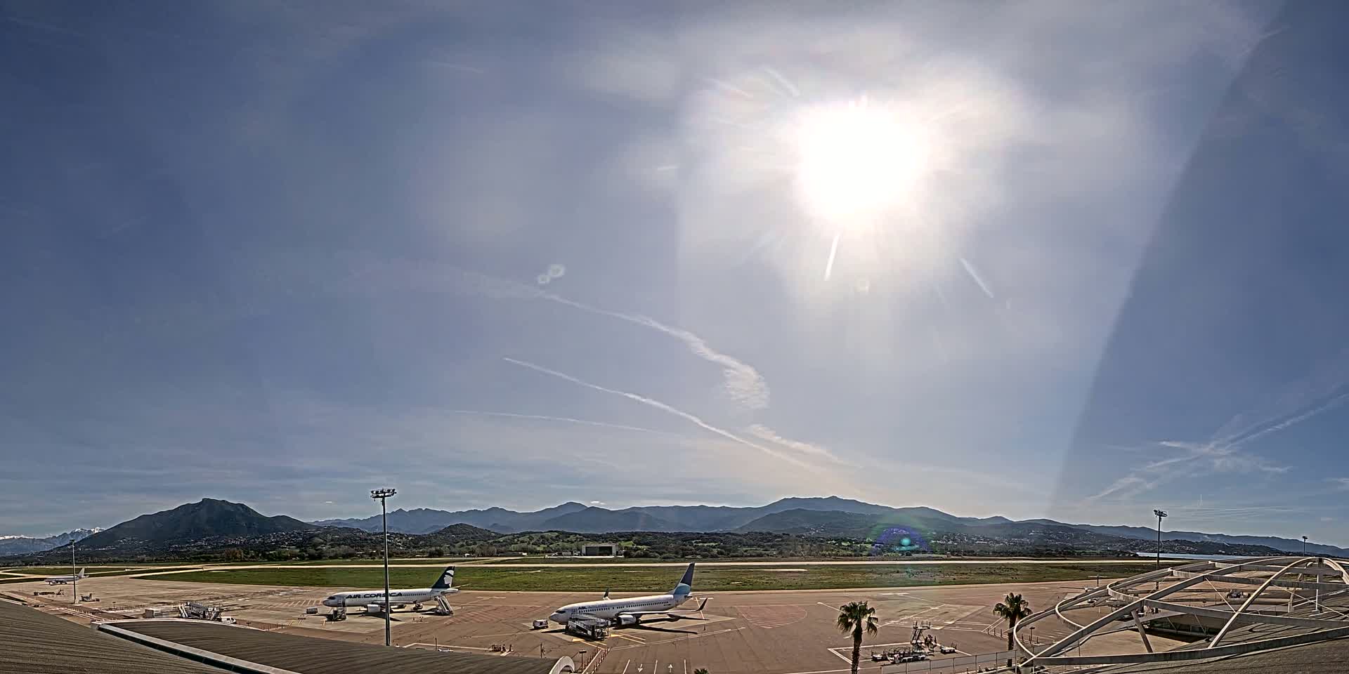 Two airplanes are parked on a sunny airport tarmac, with mountains visible in the background under a mostly clear sky.