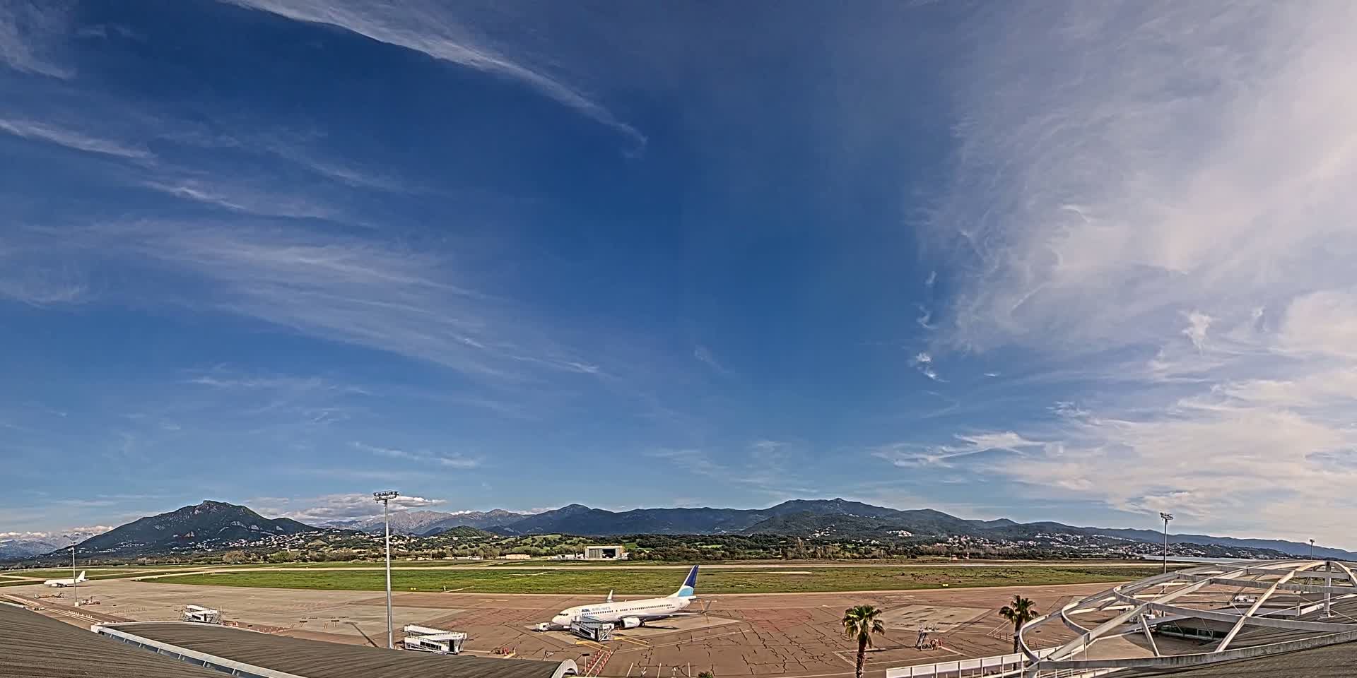 A passenger airplane sits on a tarmac at an airport under a mostly clear blue sky with wispy clouds, backed by a range of mountains.