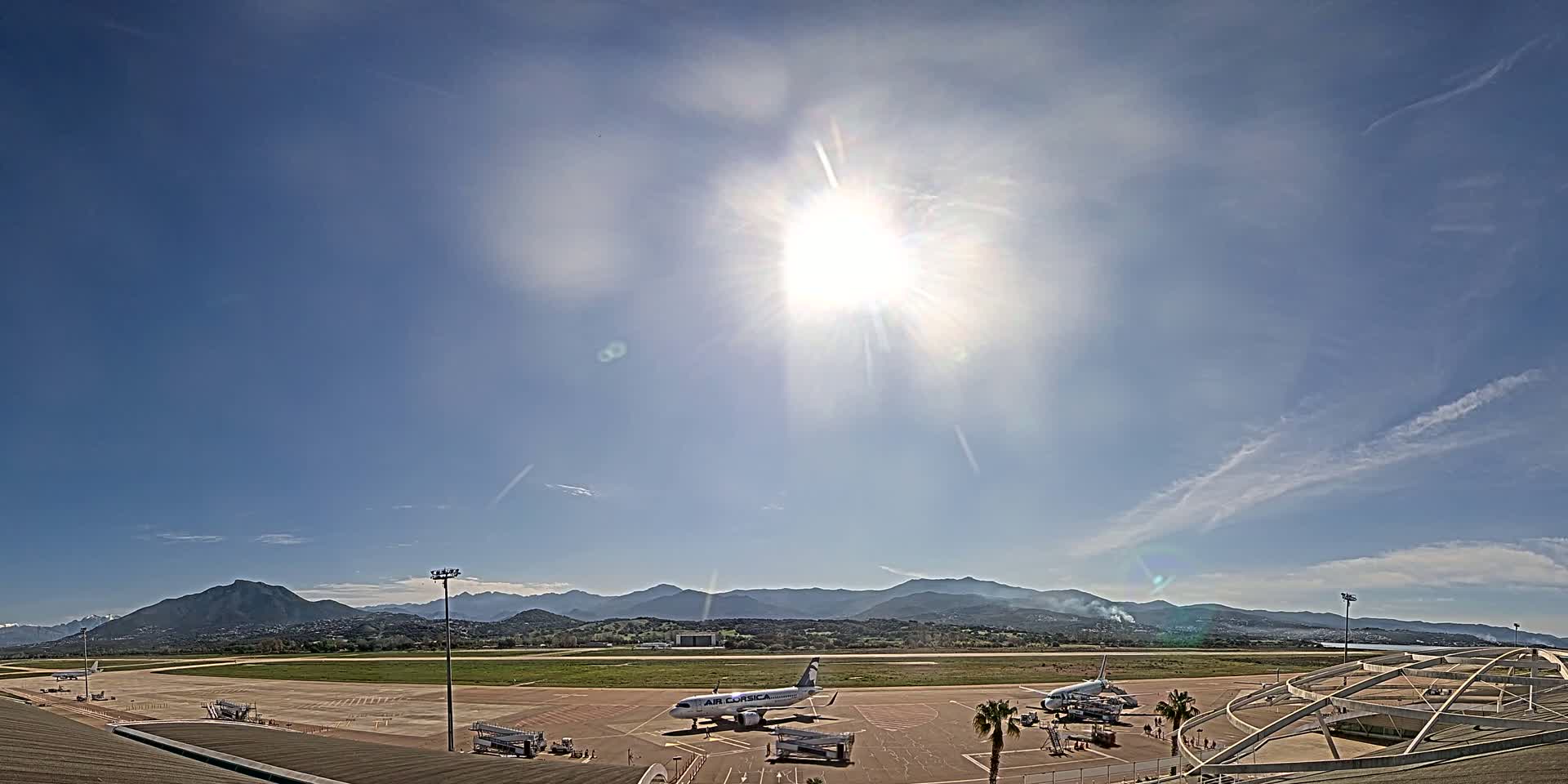 A sunny day at an airport shows several planes on the tarmac with mountains in the distance.